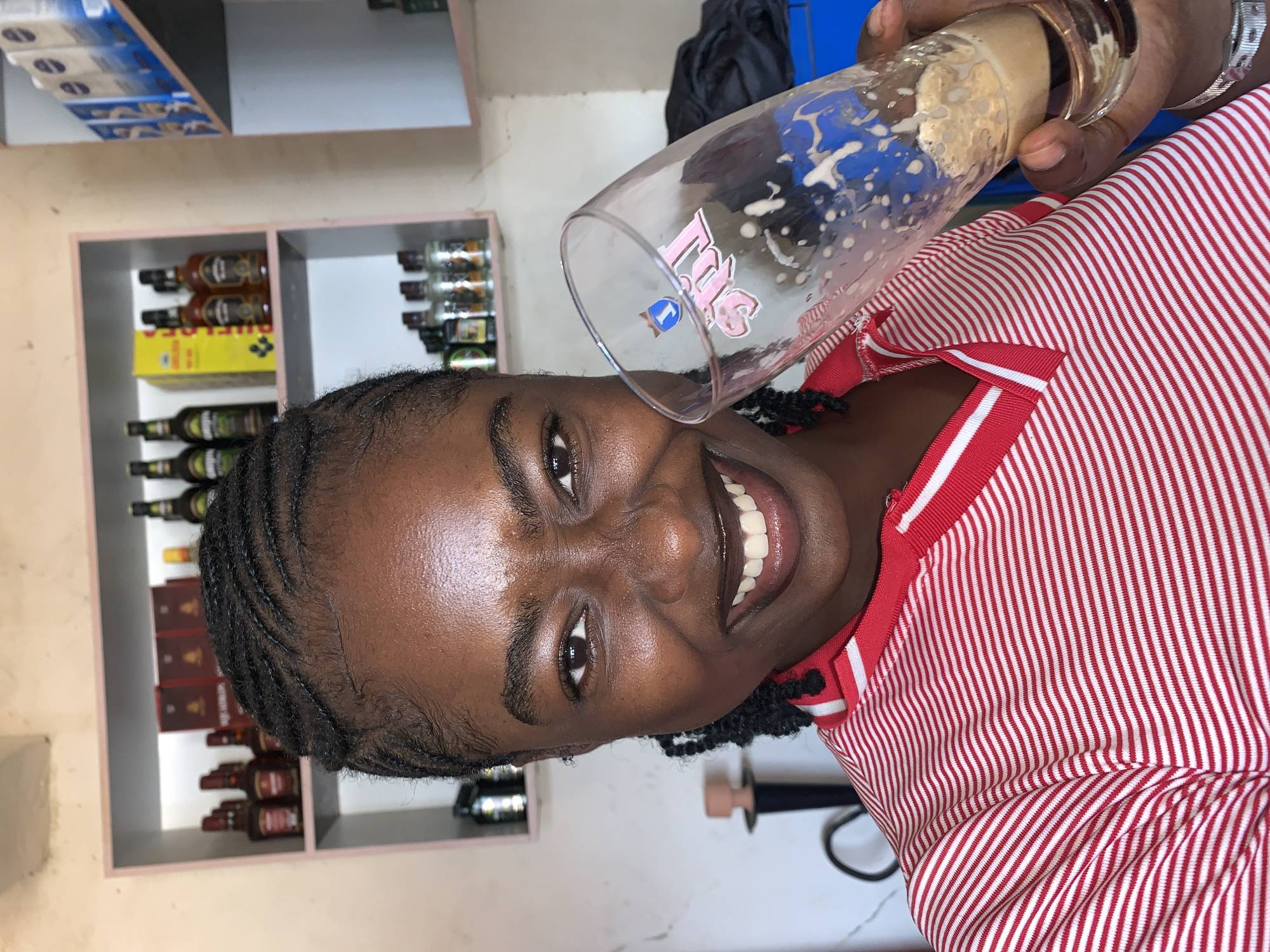 A young Black woman is captured in a close-up, waist-up shot, smiling broadly and looking directly at the camera. She has dark braided hair and is wearing a red and white striped polo shirt with a red collar and white trim. Her expression is joyful and engaging, highlighting her bright eyes and white teeth.
She holds a clear glass, typical of a beer glass, tilted upwards. The glass contains a foamy, light-brown liquid, likely beer, with some remnants of foam on the sides. Visible text on the glass includes stylized pink lettering that appears to read "Fis" or "Tis", accompanied by a blue logo that looks like a 'T' with an upward arrow or flag.
The background shows an indoor setting, likely a small shop or bar in New Stadium, Nigeria. Behind the woman, there are light-colored shelves filled with numerous bottles of various shapes and colors, suggesting different types of alcoholic beverages, such as liquor or wine. A yellow box is also seen on one shelf. Higher up, another shelf holds white packaged items. The overall scene suggests a casual atmosphere where the woman is enjoying a drink.