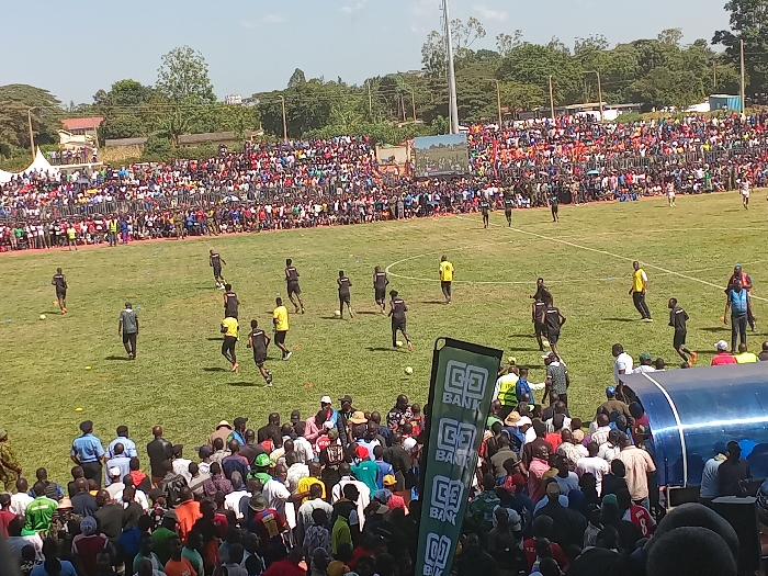 This image captures a vibrant football match or warm-up session taking place in a stadium in Siaya, Kenya, under bright, sunny daylight.
The central subject is a lush green football pitch, where numerous players are active. Most players are dressed in athletic kits, primarily black or yellow, engaged in running, passing, or drills. A football is visible on the field, confirming the sport. In the distance, parts of goalposts can be faintly discerned.
Dominating the background are densely packed stadium stands, teeming with a large crowd of spectators. Hundreds, if not thousands, of people are visible, some seated, others standing, indicating a well-attended event. The crowd's attire is varied, suggesting a diverse attendance. Beyond the stadium structures, trees and some distant buildings can be seen under a clear blue sky.
In the foreground, closer to the camera, more spectators are gathered. On the right side, a blue structure, possibly a team dugout or shelter, is visible. Adjacent to it, a tall, green vertical banner prominently displays the text "CO-OP BANK," indicating sponsorship. The bright lighting and clear conditions suggest the image was taken during the middle of a sunny day. The overall atmosphere conveys a lively community sporting event.