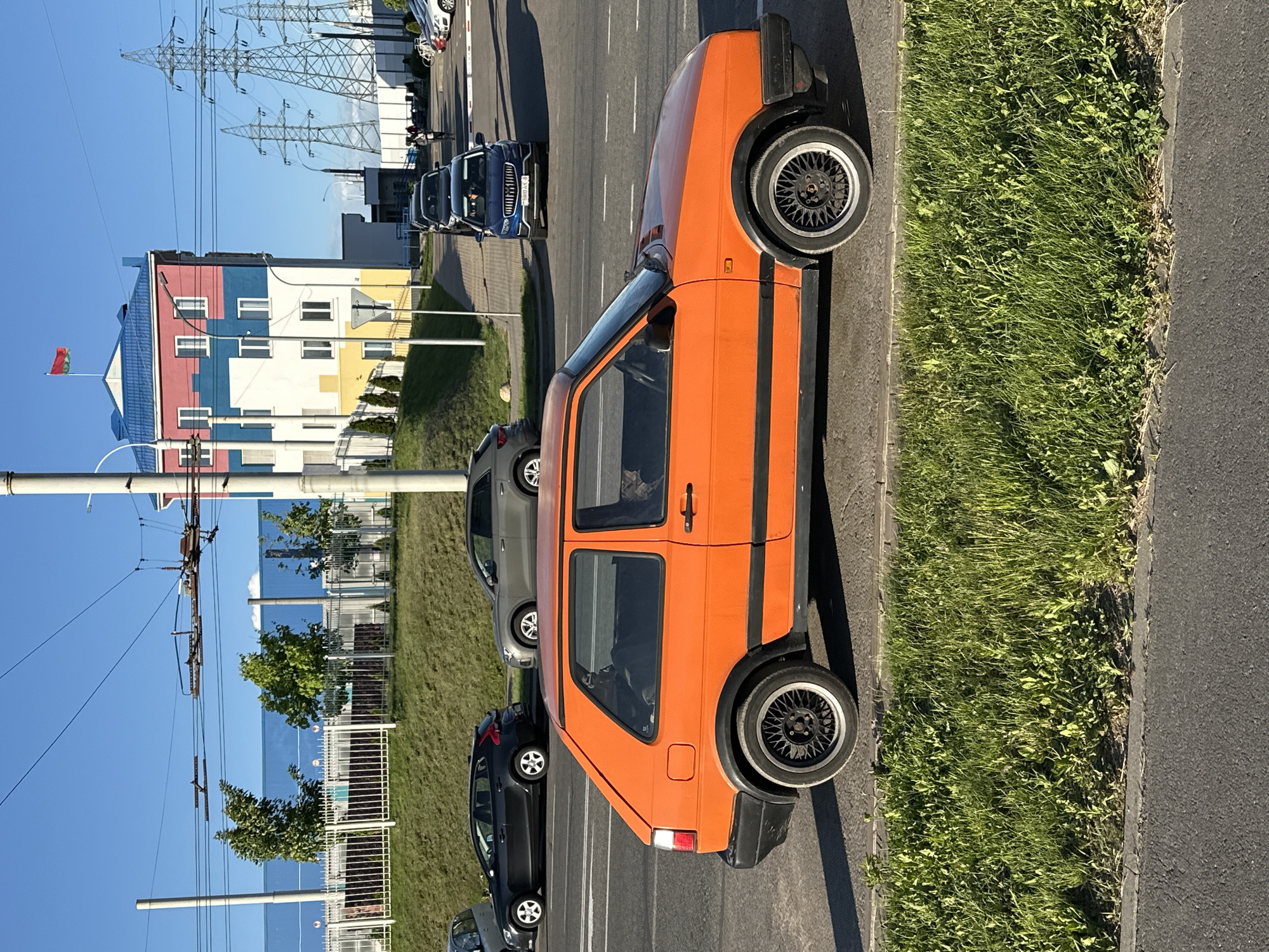 The image shows a street scene in Minsk, Belarus. An orange compact car is parked along the curb, next to a strip of grass. The road leads to a colorful building with a blue roof and walls painted in sections of red, white, blue, and yellow. A Belarusian flag flies from a pole atop the building. Overhead power lines are visible, as are the support structures for trolleybuses. Several other cars are parked further down the street. The sky is blue.