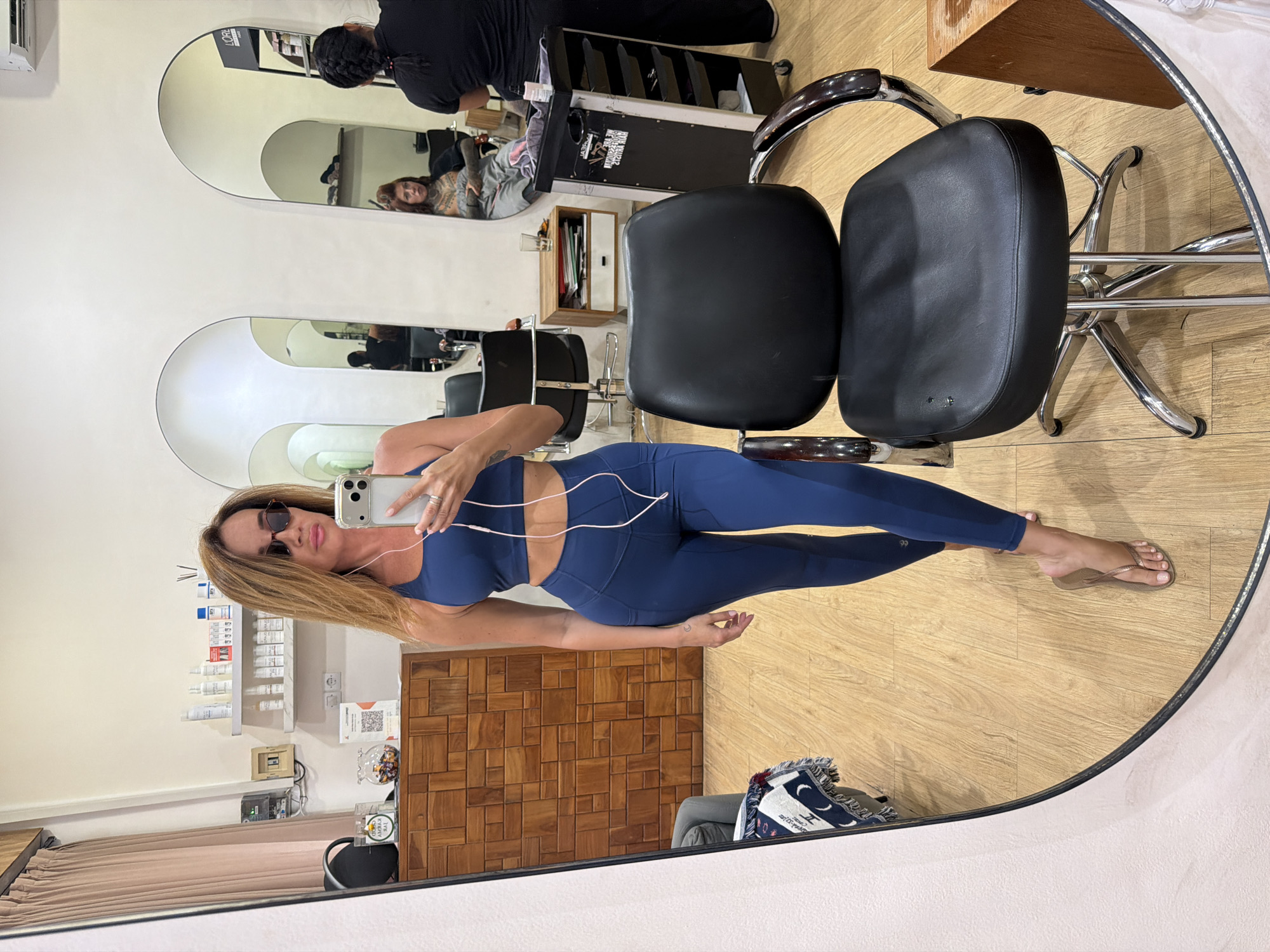 A woman stands in a salon, taking a mirror selfie. She is wearing a dark blue athletic set, consisting of a sports bra and leggings, and nude flip-flops. She has on sunglasses and is holding a smartphone in her right hand, with earbuds in her ears. The salon's interior is visible in the mirror's reflection, showing wooden cabinets, shelves stocked with products, hair salon chairs, and other salon equipment. Two women are also visible in the reflection; one is seated and appears to be a client, while another, a hairstylist, is standing and facing away from the camera. The salon floor is made of light-colored wood planks.