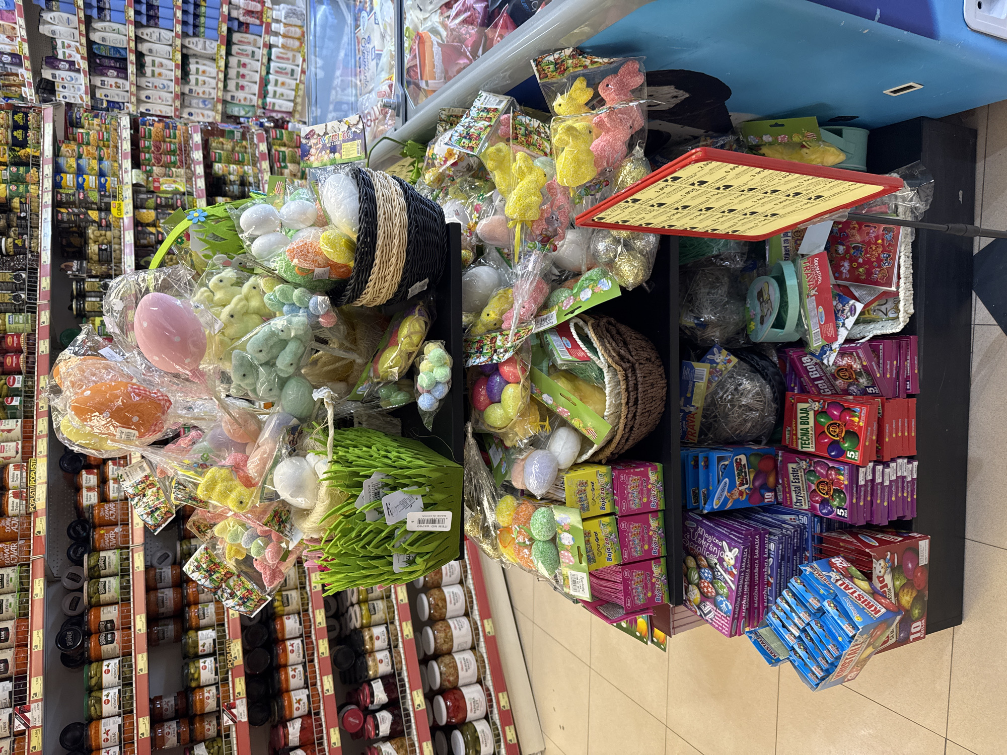 The image shows a display of Easter-themed merchandise inside a store, likely a grocery store or supermarket, in Sutomore, Montenegro. The display includes baskets filled with decorative eggs, plush chicks, and other Easter decorations wrapped in plastic. Boxes of Easter egg dye and other related items are also visible. The display is located next to shelves stocked with jars of various food products. The overall scene suggests a retail environment where Easter products are being promoted and sold.
