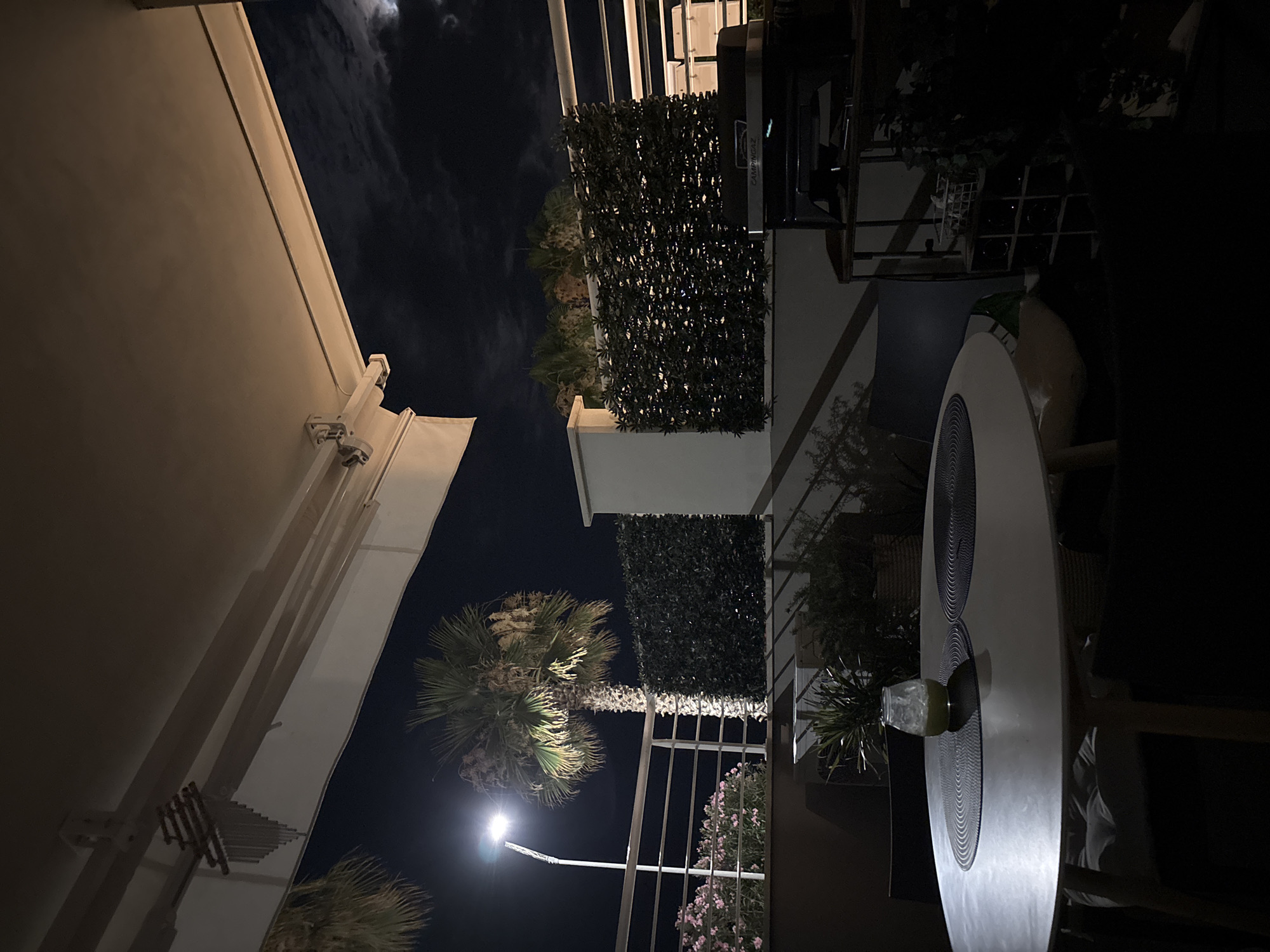 The image depicts an outdoor patio at night, likely in Cupra Marittima, Italy, as suggested by the presence of a palm tree and the overall Mediterranean aesthetic. In the foreground, a round white table is set with two dark placemats and a small glass containing a green beverage. A portion of a chair is visible to the left of the table.
The midground features a white railing, beyond which a streetlamp illuminates a tall palm tree. To the left of the palm tree, there are pink flowers. Behind the railing, a neatly trimmed hedge forms a green wall, with some of its lighting visible. To the right of the hedge, a white structure is present. Further to the right, a black Campingaz grill is situated on a shelf unit, with a white basket containing items placed on top of it. A wine rack filled with bottles is below the grill. Various plants are visible in pots and planters throughout the patio.
The background is a dark night sky, with a full moon and some clouds visible in the upper right corner. The scene is illuminated by the streetlamp, ambient moonlight, and artificial lighting from the patio itself. The overall atmosphere is serene and inviting.