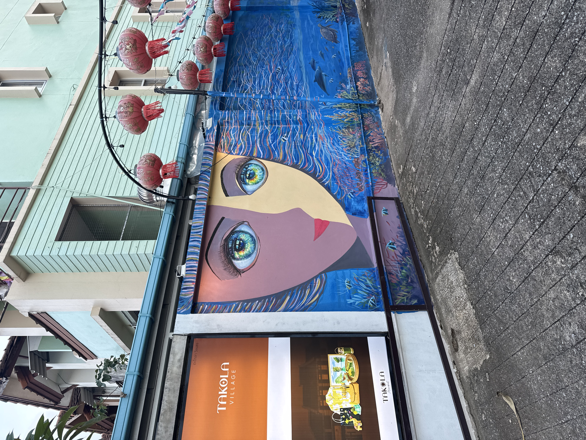 The image captures a vibrant street scene in Ban Bang Niang, Thailand. The focal point is a large mural painted on a textured wall. The mural depicts a stylized human face, split down the middle, with one half rendered in shades of purple and brown, and the other in yellow. Both halves feature striking, colorful eyes. To the right of the face, the mural transitions into an underwater scene, complete with coral reefs, fish, dolphins, and a turtle, all rendered in various shades of blue and white. Above the mural, red Chinese lanterns are suspended from a curved black wire, adding a festive touch. To the left of the mural is a large advertisement for "Takola Village," featuring images of clothing, accessories, and what appears to be a beverage or food product. The advertisement has an orange and white background. The street itself is paved with textured gray concrete, with visible lines and some patchy wear. The background shows elements of building architecture, including light blue and pale green facades with window frames. The overall impression is a colorful and lively urban environment.