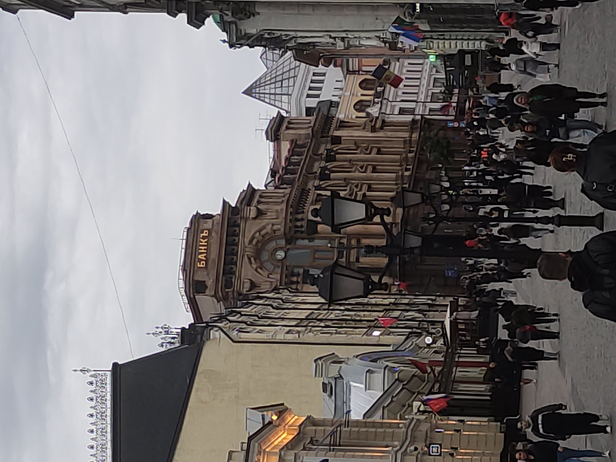 The image shows a street scene in Moscow, Russia. The street is crowded with people walking in both directions. Several buildings line the street, with one having the word "БАНКЪ" (Bank) illuminated in gold lettering on its facade. There are many flags and lamp posts along the street, and the sky is overcast.
