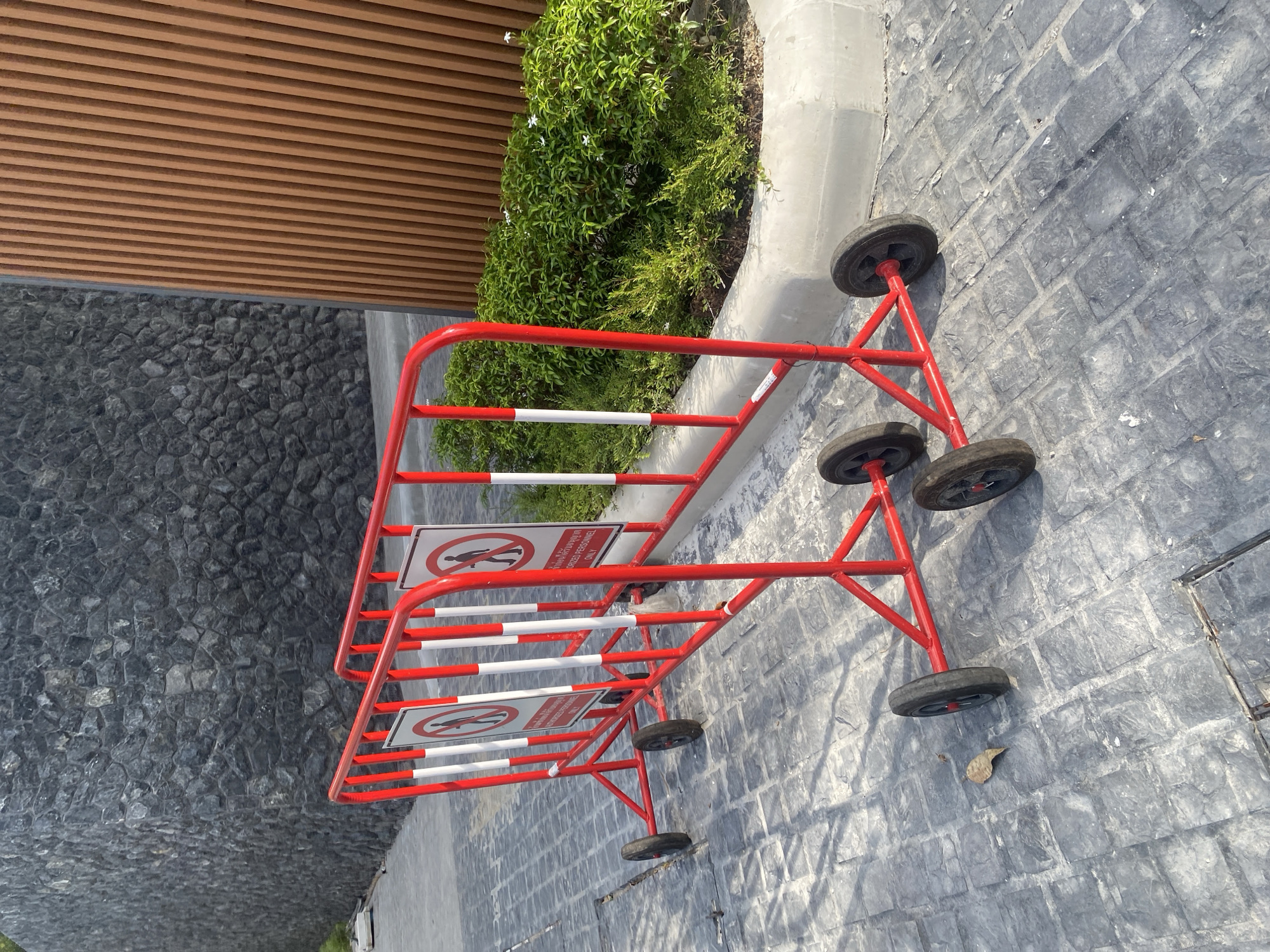 An outdoor scene in Choeng Thale, Thailand, features a red, wheeled barricade positioned on a gray, textured pavement. The barricade is marked with white stripes and signs depicting a figure with a line through it. The area is bordered by a dark gray stone wall on one side and a planter with green foliage on the other. Above the stone wall is a brown slatted structure.