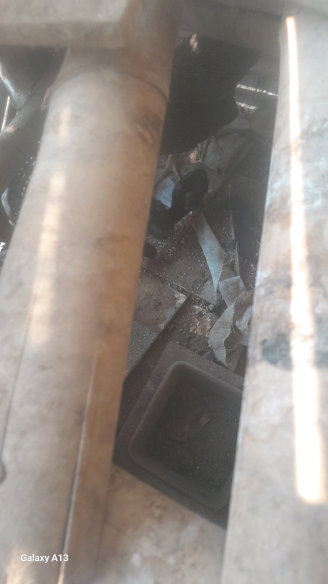 This image, taken in Hadipolo, Indonesia, shows a close-up view of pipes and debris. A large, cylindrical pipe, possibly made of metal, is prominently featured in the foreground, obscuring much of the background. The pipe appears weathered and possibly rusty.
Behind the pipe, the scene is cluttered with various objects and materials. There are pieces of what looks like rolled-up fabric or sheeting, exhibiting a textured surface and light coloring, spread out on the ground. The ground itself is covered in dirt, small stones, and possibly other granular material. A dark, rectangular container or trough is visible, partially filled with dirt and debris. Shadows suggest this is an outdoor or dimly lit area. The overall impression is one of disarray and neglect, possibly a workshop, storage area, or an area undergoing construction or demolition. The "Galaxy A13" watermark at the bottom left indicates the device used for the photograph.