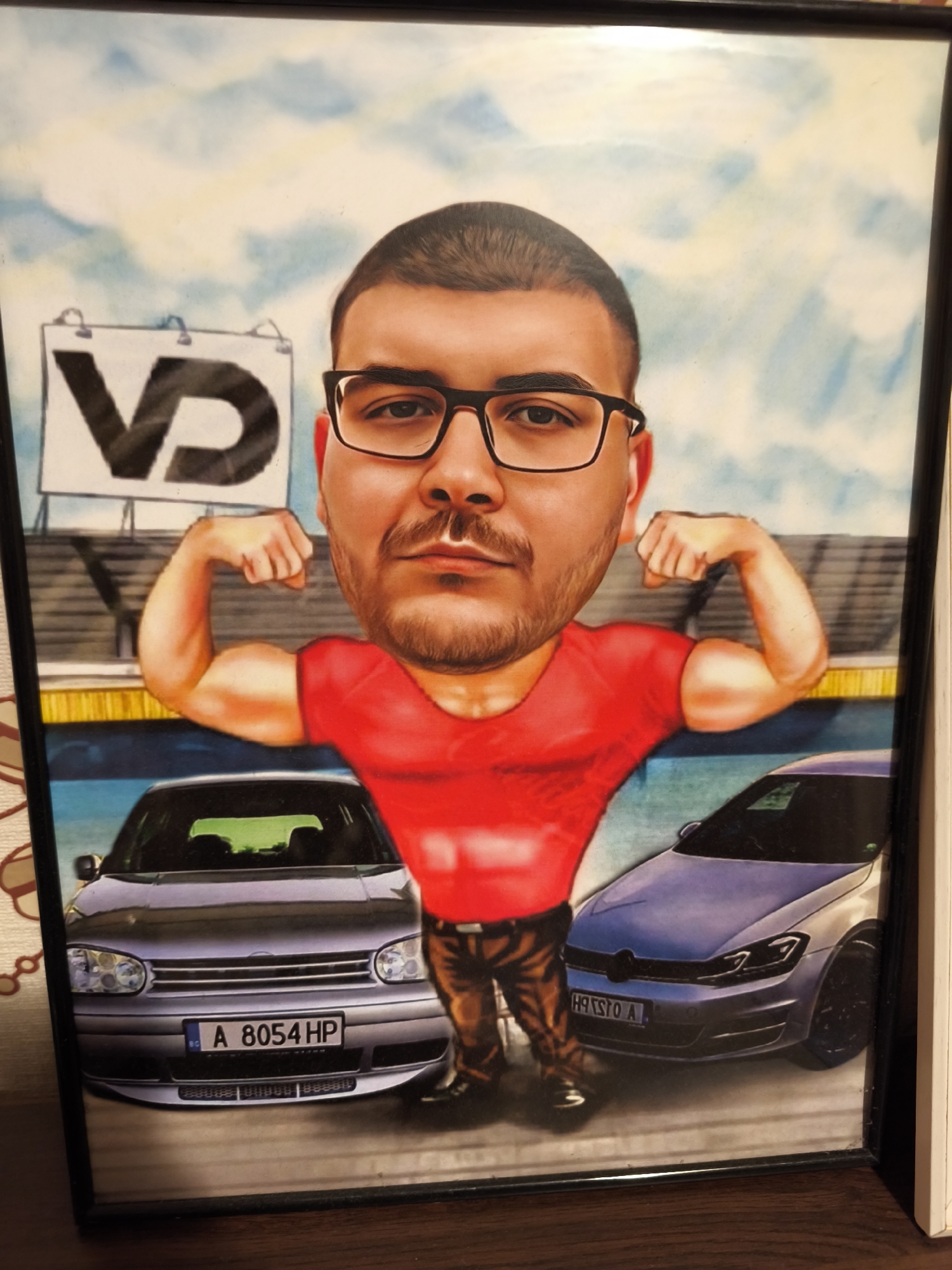 The image is a framed caricature depicting a man with an enlarged head, glasses, and a red t-shirt, flexing his biceps. He stands between two cars: on the left is an older model Volkswagen Golf with the Bulgarian license plate "A 8054 HP," and on the right, a newer Golf model with license plate "H 91570 A." Behind the man and cars is a backdrop that includes stadium seating and a sign with the letters "VD." The backdrop's sky suggests an outdoor location. The caricature is in a black frame, resting on a brown wooden surface. The license plates indicate that the scene is meant to represent Burgas, Bulgaria.