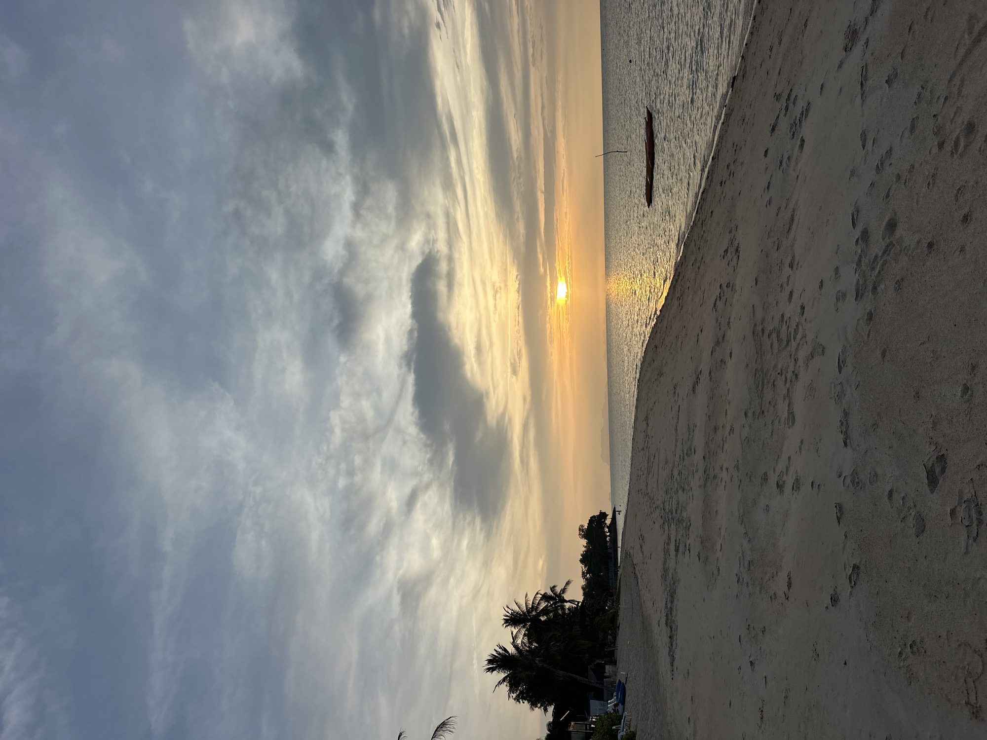 The image depicts a serene beach scene in the Ko Samui District of Thailand during what appears to be sunset or sunrise. A sandy beach occupies the foreground, marked by numerous footprints. The water of the sea is calm, with a gentle ripple on its surface. In the distance, two small boats are anchored, one of which has a pole sticking out of it. The shoreline is lined with palm trees and vegetation, casting shadows on the sand. Further back, structures that appear to be bungalows or resort buildings are visible among the trees. The sky is filled with dramatic clouds, ranging from dark gray to lighter shades, with the sun peeking through, casting a warm, golden light across the horizon and reflecting on the water. The overall atmosphere is tranquil and picturesque.