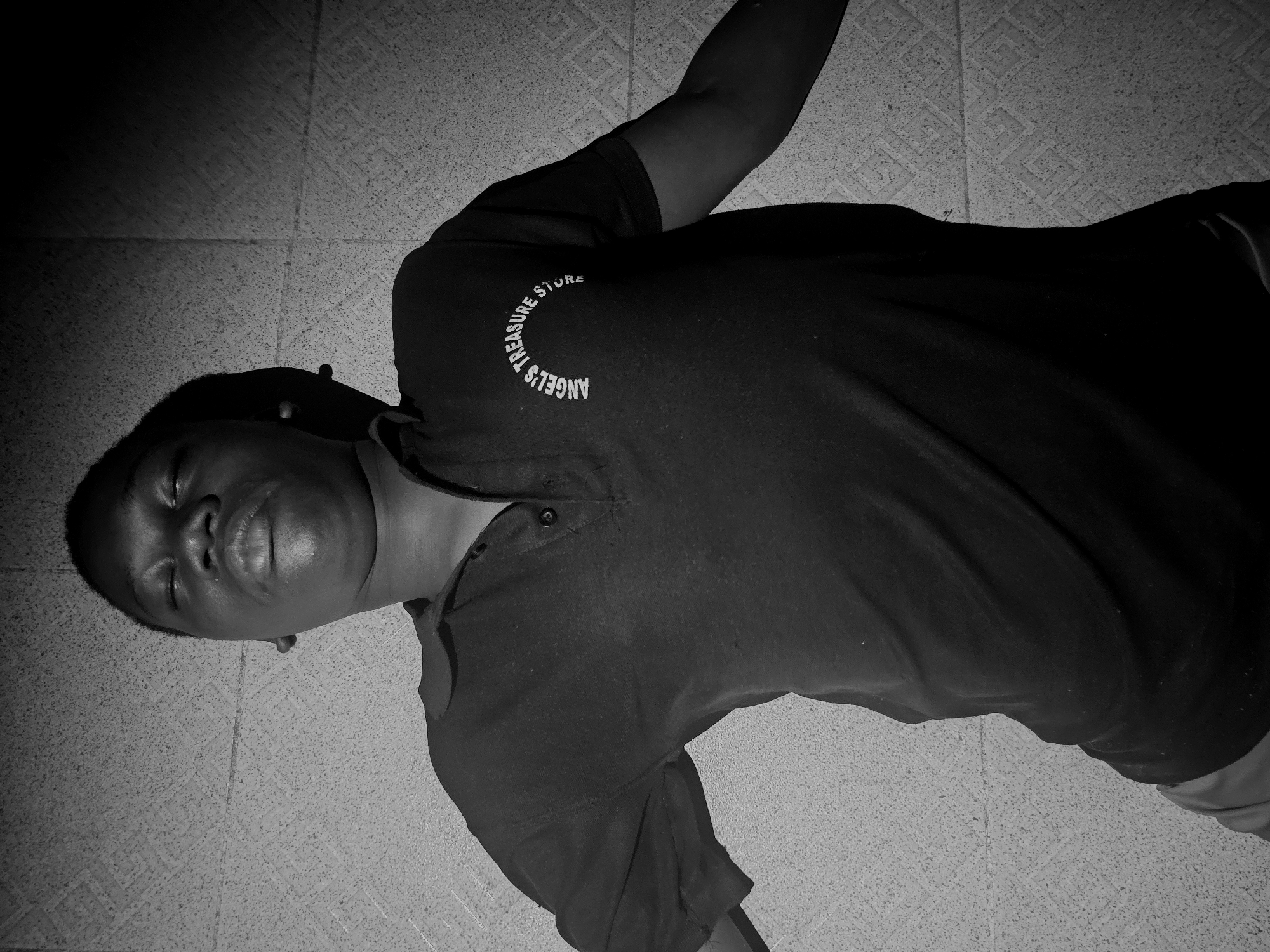 A medium shot, taken from a low angle, shows a Black man lying on his back on a tiled floor in a location within the city of Asaba, Nigeria. The man is wearing a dark t-shirt with "ANGEL'S TREASURE STORE" printed in a circular pattern. His left arm is extended above his head. His eyes are closed, and his facial expression is relaxed. The patterned tile floor is visible beneath him.
