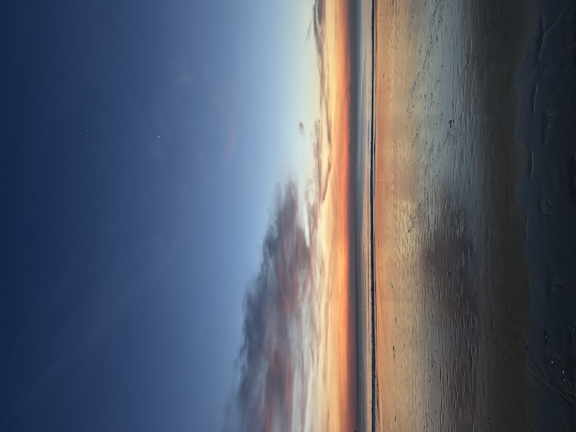 The image depicts a sunset at a beach in Kuta Selatan, Indonesia. The sky transitions from a dark blue to lighter blue with scattered clouds near the horizon, where a gradient of orange and yellow hues marks the sunset. The beach sand is wet, reflecting the colors of the sky, and it becomes darker towards the foreground. The horizon line is thin, with a small visible ocean band.