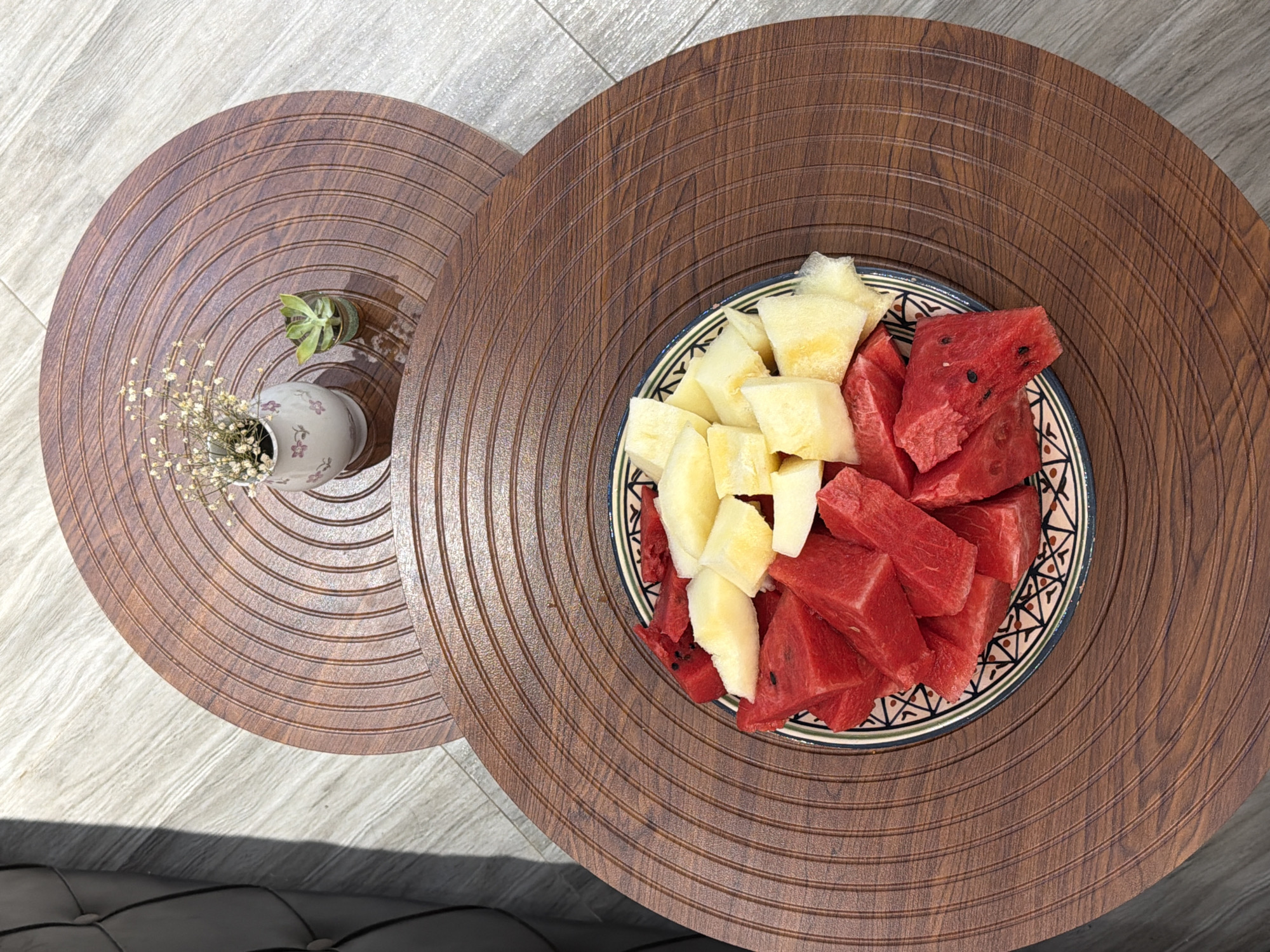 The image shows a close-up, overhead view of a fruit platter on a wooden table. The platter is filled with cubed watermelon and pineapple. The table is circular with a wood-grain pattern and concentric rings. In the background, another smaller circular table with similar detailing holds a small vase with dried flowers and a succulent plant. The floor appears to be tiled. The image does not provide any information about the city of Sabirabad, Azerbaijan, or any people or actions.