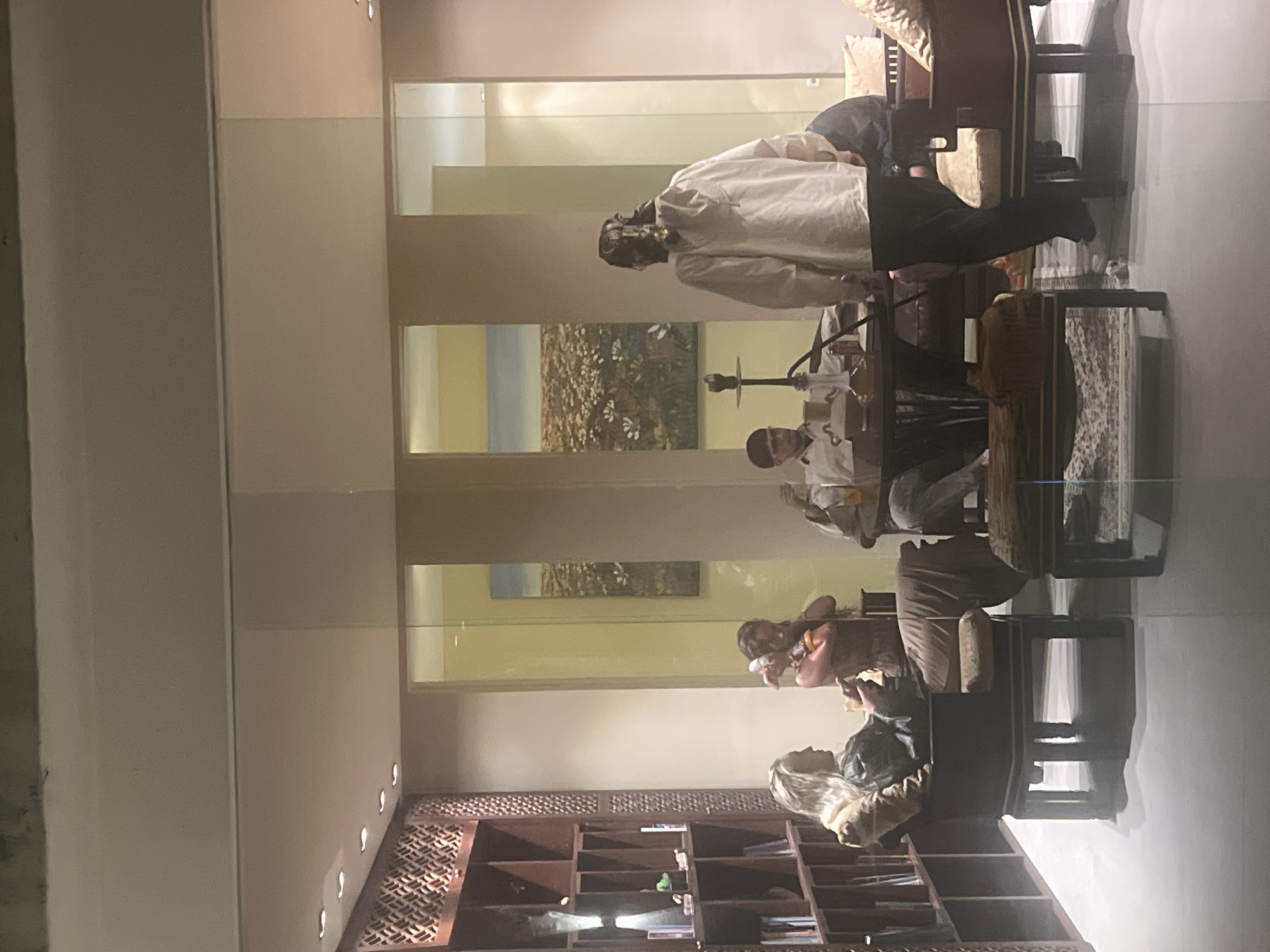 This image captures an indoor scene within a lounge or common area in Kuta Selatan, Indonesia, viewed through a glass partition. Five individuals are present in a furnished space featuring modern and traditional elements.
On the far left, a person with long blonde hair is seated in a dark wooden armchair, facing right, looking down at an object held in their hands. They wear a dark jacket over a lighter garment and light-colored trousers. Adjacent to them, a decorative dark wooden shelving unit with geometric lattice patterns fills the left wall, containing books and various small objects.
In the center-left, a person with dark hair is standing, facing slightly left, holding what appears to be a small child or a wrapped object close to their chest. They are dressed in a patterned, dark-toned top.
At a low, round dark wooden table in the center, a person with dark hair and glasses is seated, facing left. This individual wears a light-colored long-sleeved shirt with a visible "CUP" logo and holds a drink in their right hand. A prominent hookah (shisha pipe) with a clear glass base and dark hose rests on the table in front of them, along with another glass.
Behind the central table, a person with long dark hair tied up, wearing a light-colored hooded jacket and dark pants, stands with their back mostly to the viewer, facing towards the right side of the room. Their left foot is slightly elevated.
To the far right, a fifth person is partially visible, seated on a dark sofa with a light-colored cushion and a patterned pillow.
The room features light-colored walls, possibly beige or cream, with sections divided by pillars and glass panels. Through these glass panels, two identical or very similar landscape paintings depicting a field of white flowers under a blue sky are visible on the back wall. The floor is a light grey or white tile. Recessed lighting illuminates the ceiling on the left side of the room. A patterned rug is visible beneath the central table and a dark wooden footrest in the foreground. Reflections from the external environment are apparent on the glass through which the photograph was taken.