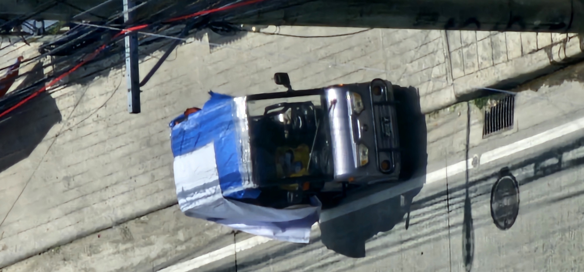 This is a top-down, slightly angled, medium-resolution image of a small, three-wheeled vehicle covered with a blue and white tarp, parked on a concrete road. The vehicle appears to be a type of autorickshaw or "tricycle" commonly used for transportation in the Philippines. The vehicle has a silver body, a metal bumper with circular orange reflectors, and a license plate with illegible characters. The driver's cabin has windows and side mirrors. The tarp is draped over the roof and sides of the vehicle, with the blue and white pattern visible.
The vehicle is positioned on the right side of a two-lane road, indicated by white lane markers. To the left of the vehicle, a manhole cover is visible on the road surface, along with a storm drain grate. The road appears to be made of worn concrete, with some cracks and patches. A sidewalk with a tiled surface is visible to the right of the road. Overhead, a dense network of electrical wires and cables is suspended, supported by a utility pole on the right side of the frame. Shadows suggest it is daytime.
The image quality is affected by compression and blurriness, making fine details difficult to discern. Based on the presence of the utility pole with dense wiring, the style of the vehicle, and the road infrastructure, this scene is consistent with an urban environment in the Philippines. Specific details for the city of Makati, Philippines are not discernible from the visual information present in the image.