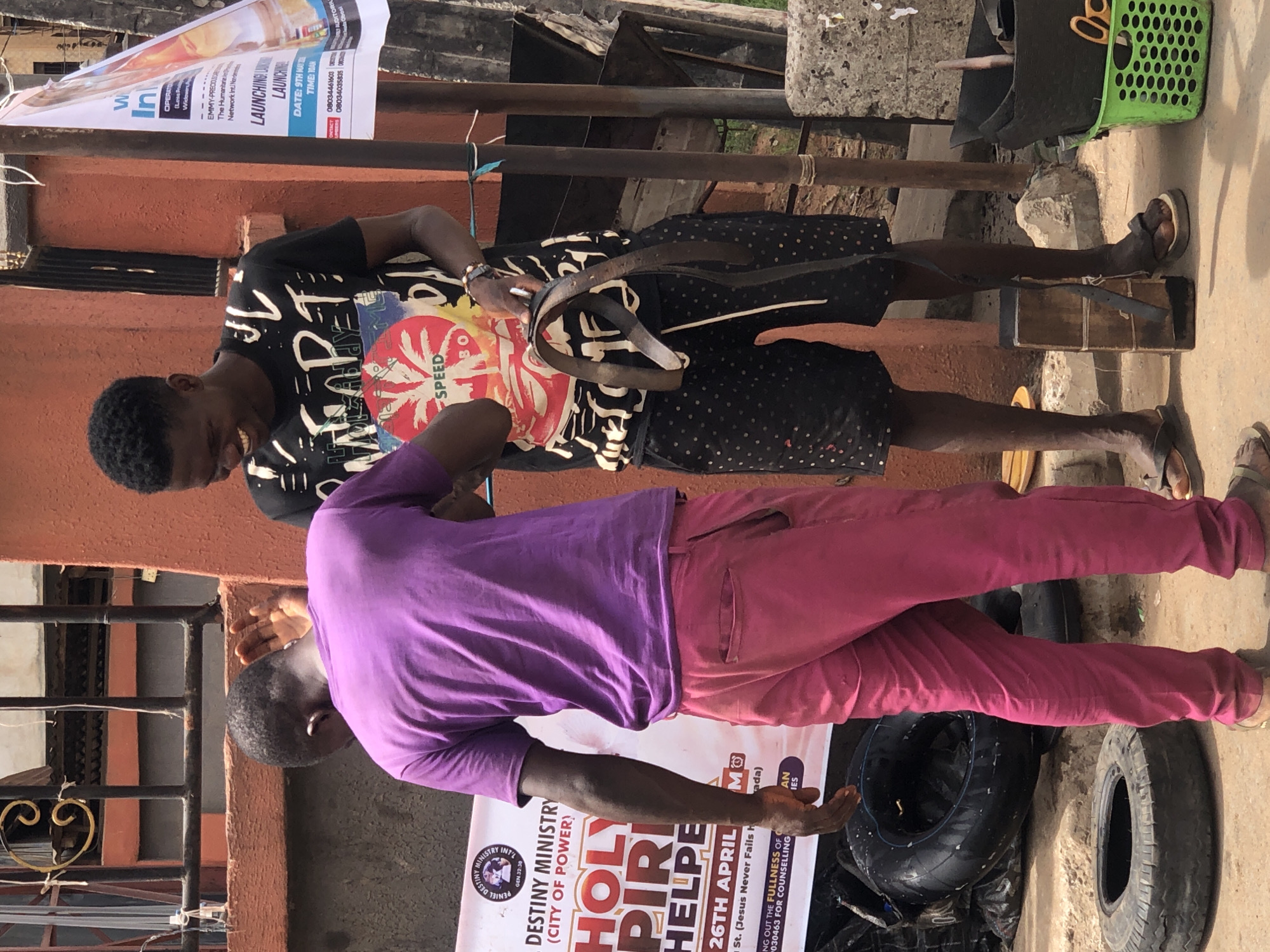 The image shows two African men standing outdoors. One man is wearing a purple shirt and pink pants. The other man is wearing a black t-shirt with a design, and black shorts with white dots. Both men are dark-skinned. A green basket, a box, and tires are visible. Banners or signs are posted. One sign mentions “Destiny Ministry” and “Holy Spirit Helpers” with a date. Another advertises a launching event. The OCR implies that the scene is in the city of Onitsha, Nigeria.