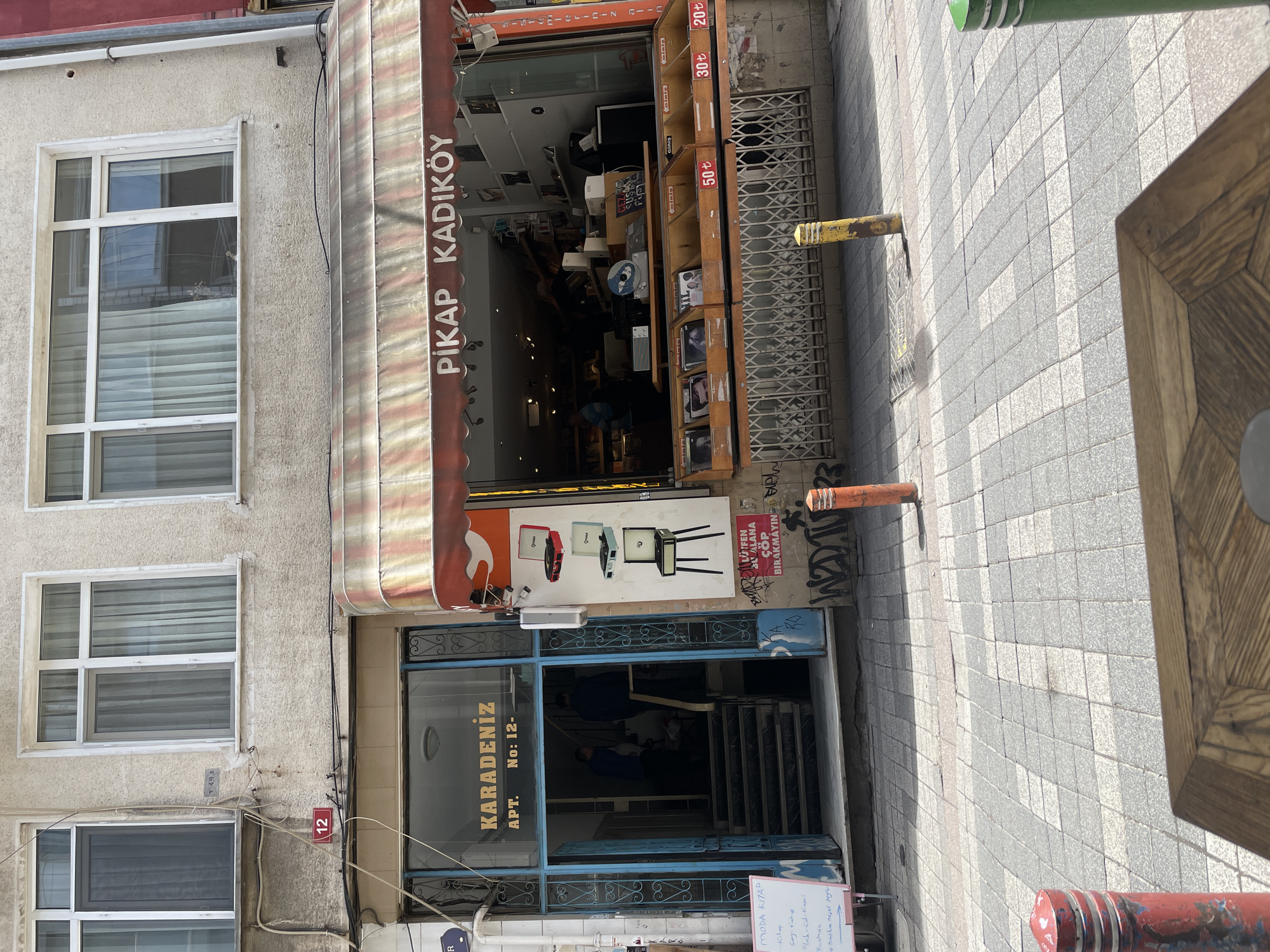 The photograph presents the street-level view of two businesses located next to each other in the Kadıköy district of Istanbul, Turkey. On the right, a record store named "Pikap Kadıköy" is visible, identified by its awning. The store displays record covers and pricing on shelves extending to the sidewalk. Next to "Pikap Kadıköy" is an entrance to "Karadeniz Apt. No: 12," a building with a blue-painted door and a sign bearing the building name and address. A staircase leads up from the street into the building. A white board near the entrance has hand written text which includes “MODA KITAP” at the top and underneath are the words Kitap, Gay-Kabe, Plab-cd-Kase, Baha, ve basla nejel segle. The structure to the left of the building entrance has three window sets that appear to be a residence. A “12” street number can be seen below the bottom window. A table sits on the right side of the photograph and on the sidewalk are two posts.
