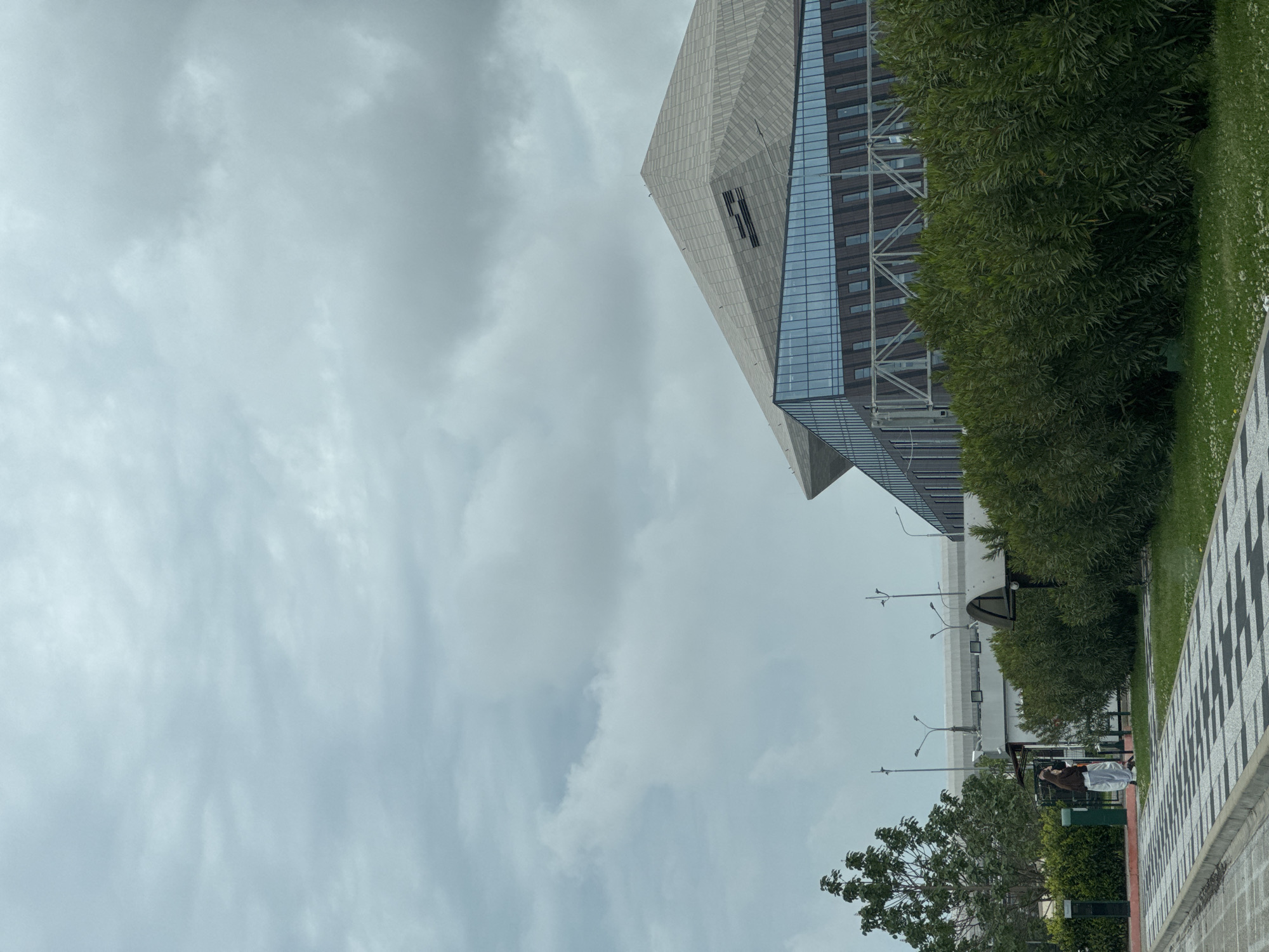 The image depicts a cloudy sky above a modern building in Sochi, Russia. The building features a gray, angular design with a glassy facade. A tree and green vegetation fill the foreground to the right. A sidewalk runs along the bottom of the image, with streetlights visible in the background. In the bottom right, there appears to be a person sitting on a bench. The day appears overcast.