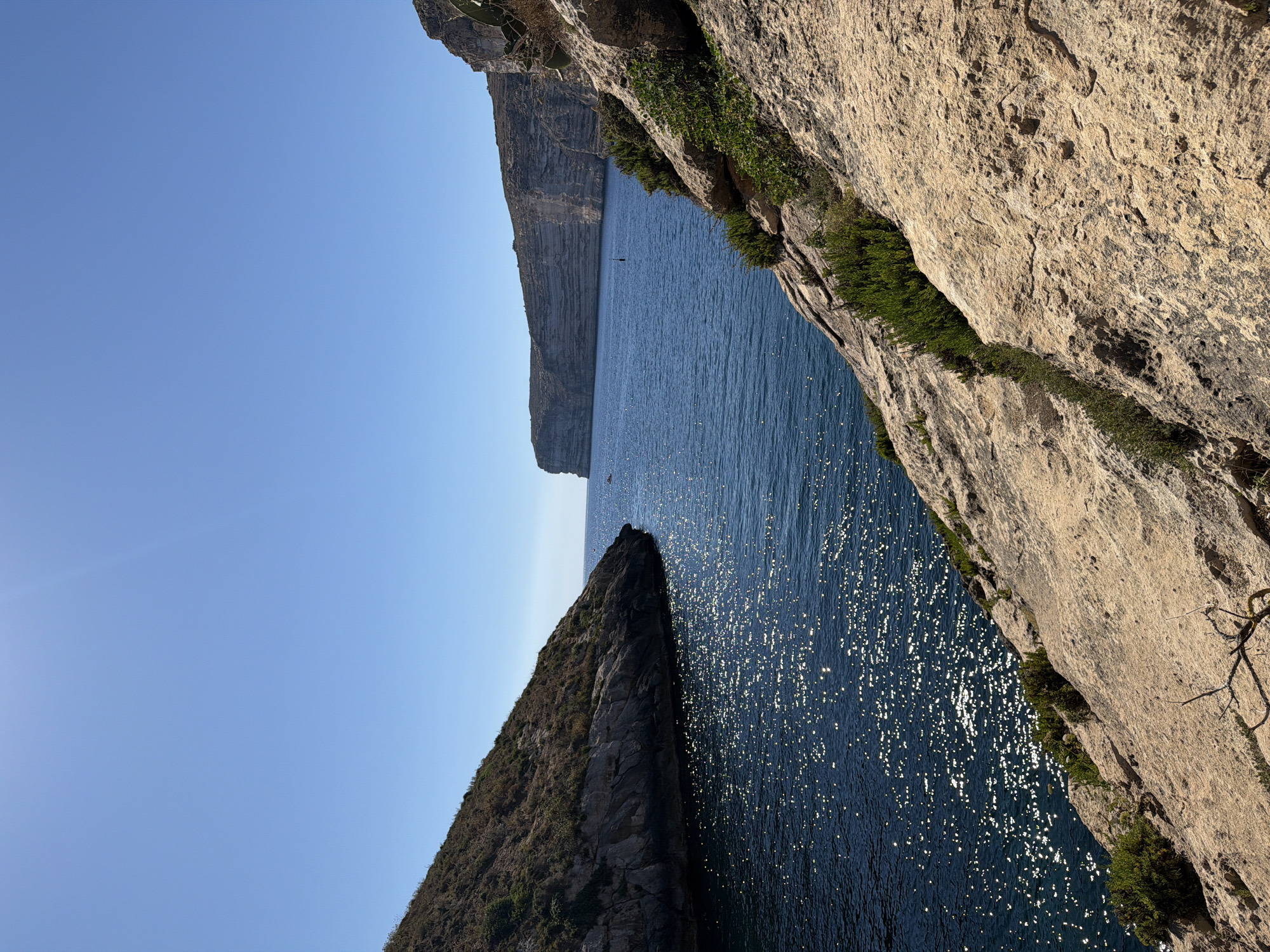 This image depicts a picturesque coastal scene in Xlendi, Malta. The foreground is dominated by a rocky incline with sparse vegetation, leading down to the deep blue water of the Mediterranean Sea. The sea is calm, with sunlight glinting off its surface, creating a shimmering effect. In the middle ground, two prominent rock formations jut out into the sea, creating a bay. The larger formation on the left is covered in dry vegetation, while the formation on the right is more rugged and rocky. In the distance, dramatic cliffs rise from the sea, forming a natural barrier. The sky is a clear, bright blue, with no clouds visible. A small boat and a buoy are visible in the water.