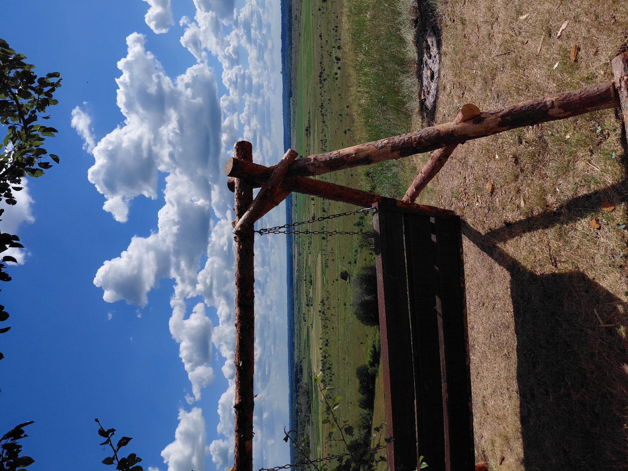 A handmade wooden swing with a dark wooden seat is positioned on a grassy knoll overlooking a vast, green landscape. The sky above is a vibrant blue, dotted with fluffy white cumulus clouds. The swing is constructed from rough-hewn logs and supported by sturdy wooden posts. Chains connect the swing seat to a horizontal wooden beam. In the foreground, dry grass and scattered leaves cover the ground. The background reveals a sprawling vista of fields and trees stretching to the horizon, under a bright, sunny sky. To the right of the swing, a circular area of dark ash suggests a recent campfire. The image appears to be taken in Ustyvytsia, Ukraine, on a clear day, offering a tranquil, natural setting.