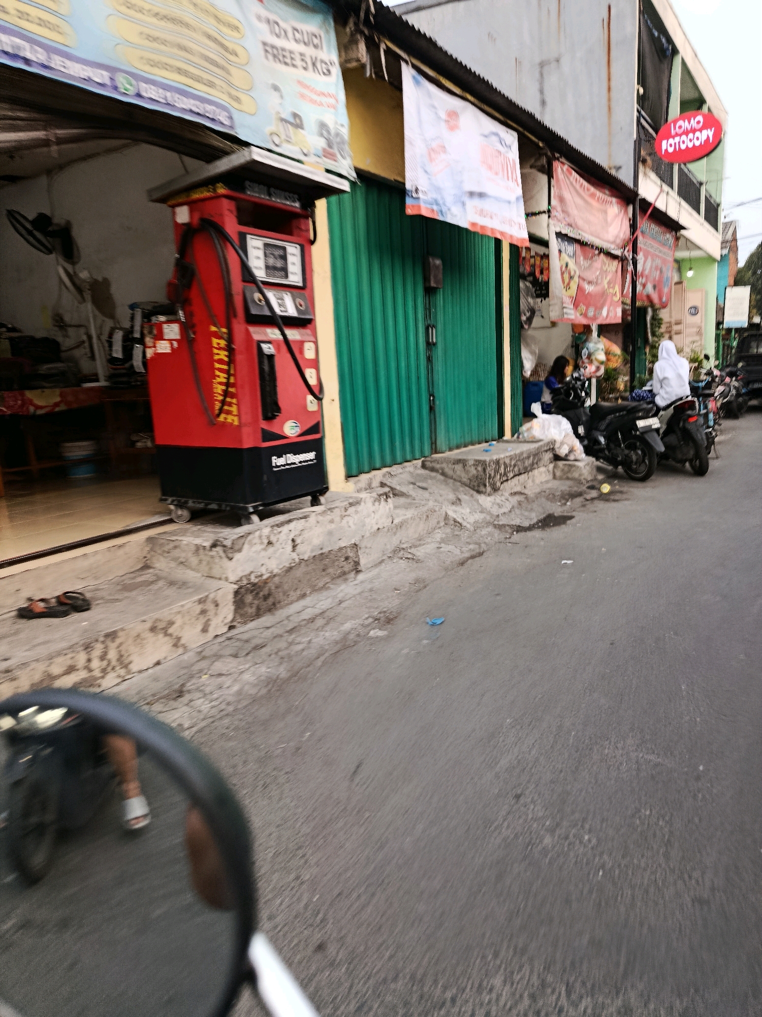 This is a street-level, eye-level view captured in Bekasi, Indonesia. The image depicts a roadside scene with several commercial establishments. In the foreground, the asphalt road is visible, with some debris, including plastic bags, scattered on its surface. To the left, a raised concrete structure with steps leads up to a storefront. On these steps, a pair of flip-flops is placed.
A prominent red and black fuel dispenser stands on the side of the road, equipped with hoses and a digital display, indicating it is a "Fuel Dispenser" and likely an "EXTREME" brand. The storefront behind it has a green metal roll-up door. Above the storefront, banners and signs are displayed, one of which reads "LOMO FOTOCOPY" in red lettering within a circular sign.
Further down the street, several motorcycles are parked. Two individuals are visible, one seated on a motorcycle and another standing near it, both appearing to be engaged in casual interaction. The building facades suggest a busy urban environment, typical of a commercial area in Bekasi. The overall lighting suggests it might be late afternoon or early evening, with overcast skies contributing to a diffused light.