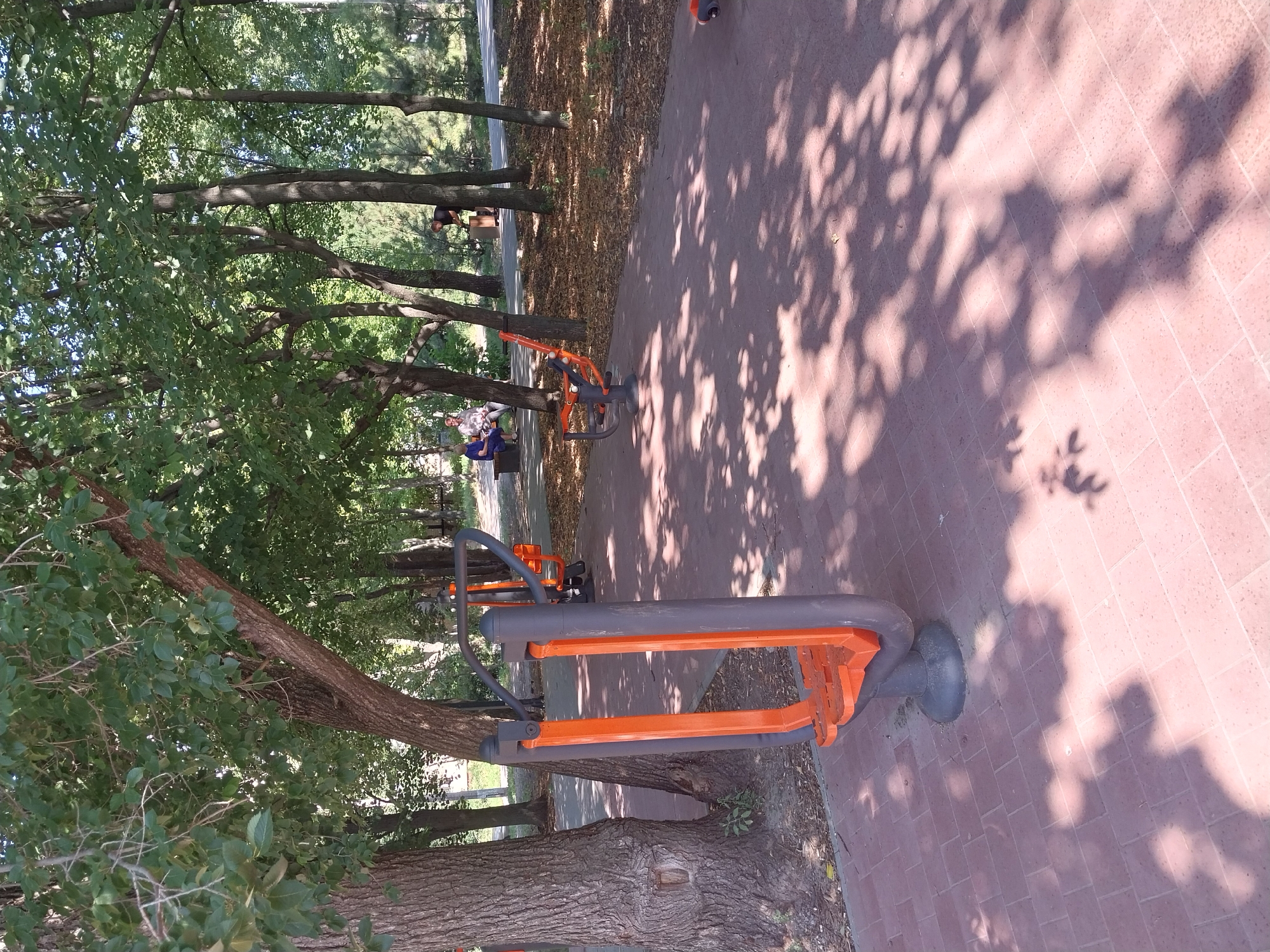 The image depicts an outdoor fitness area in a park. In the foreground, a street workout machine with orange and gray components is visible. This machine appears to be designed for leg exercises. The ground is paved with red brick-like tiles, and dappled sunlight filters through the dense green canopy of mature trees. In the middle ground, another similar exercise machine is present. Further in the background, two individuals are seated on a park bench, and another person is standing near a wooden structure. The environment suggests a public park setting, likely in a city, as indicated by the presence of well-maintained public amenities. The trees, paving, and fitness equipment are typical of urban green spaces designed for recreation. While the provided crops offer closer views of specific elements, the overall scene suggests a well-lit outdoor environment on a clear day. There are no explicit visual cues within the image itself to confirm the specific location as Dnipro, Ukraine, beyond the user's prompt.