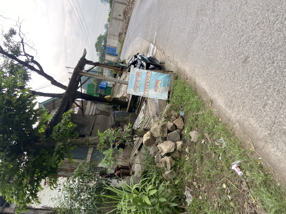 The image shows a motorcycle parked on the side of a paved road in front of a small building. The building is partially obscured by a large tree and a wooden awning. A person is visible sitting on a concrete step, wearing a blue shirt and black pants. A pile of rocks sits next to the person and motorcycle, indicating a possible construction site. A blue and white sign with the words "Cuci Motor ZM" stands beside the motorcycle. The sign is likely for a nearby motorcycle wash. The road is empty and the sky is overcast with a few white clouds. The scene is located on the side of a small, residential road in the Kecamatan Margaasih district, Indonesia, with the GPS coordinates of latitude: -6.964, longitude: 107.539. The image was taken from a perspective slightly above ground level. There is a single empty plastic cup laying on the ground.