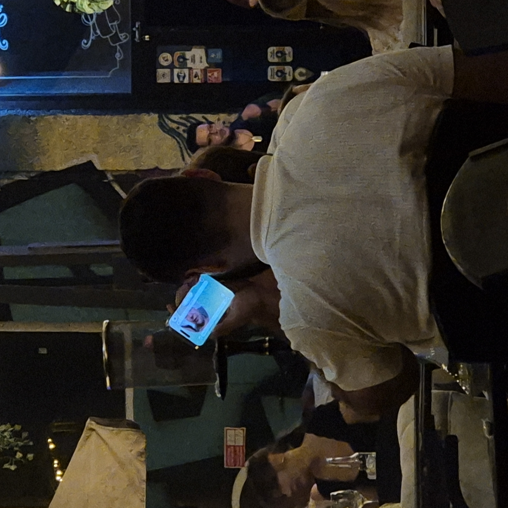 The image depicts an outdoor nighttime scene at a venue, possibly a restaurant or bar, which is located in Kemerovo, Russia. In the foreground, a man wearing a light-colored, textured polo shirt is shown from behind, holding up a smartphone that displays a brightly lit image of a person's face. The phone is held at an angle, suggesting the man is showing the screen to someone or taking a picture/video.
To the left of the man in the foreground, a woman with dark hair, wearing a black top, is partially visible. She appears to be seated at a table, with a wine glass and a bottle on it. Further to the left, a portion of an outdoor umbrella with string lights is visible, indicating an outdoor seating area.
In the background, another man with glasses and a dark tank top is visible, wearing a pendant necklace. He is also holding something in his hand, but it is unclear what it is. Behind him, a dark wall is decorated with various stickers or small posters. The overall ambiance suggests a social gathering in a dimly lit, urban outdoor environment. The quality of the image is somewhat grainy, typical of low-light photography.