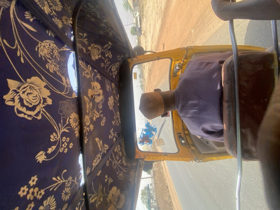 The image captures the interior of a yellow three-wheeled vehicle, likely a motorized rickshaw, in Baganakwo, Nigeria, viewed from the perspective of a rear passenger looking forward. The vehicle's roof is lined with dark blue fabric adorned with a repeating pattern of light-colored floral and foliage motifs; a small tear is visible in the fabric on the upper right side.
A single male individual is seated in the driver's position, viewed from behind. He wears a light purple, possibly striped, long-sleeved garment and a light brown, mesh-textured kufi-style cap. Blue plastic decorative items hang from the rearview mirror, visible through the windshield.
The vehicle is in motion on a paved asphalt road, which appears to be a two-lane thoroughfare. The roadside consists of dry, reddish-brown earth with sparse, desiccated vegetation. In the distance on the left, a structure featuring a green and white canopy is visible, consistent with a fuel station. On the right side, further from the road, indistinct buildings and a white truck are discernible against a clear, bright sky.
