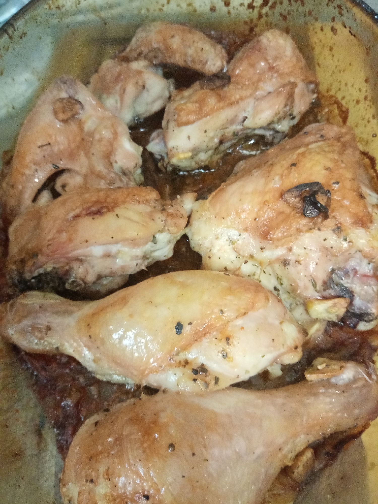 The image shows a close-up of cooked chicken pieces in a baking dish. There are several chicken pieces including drumsticks and other cuts with skin on. The chicken is lightly browned and appears to be seasoned with herbs. There is some liquid in the dish, likely cooking juices. The baking dish is a metal or ceramic pan with visible wear. There is no indication of location or city.