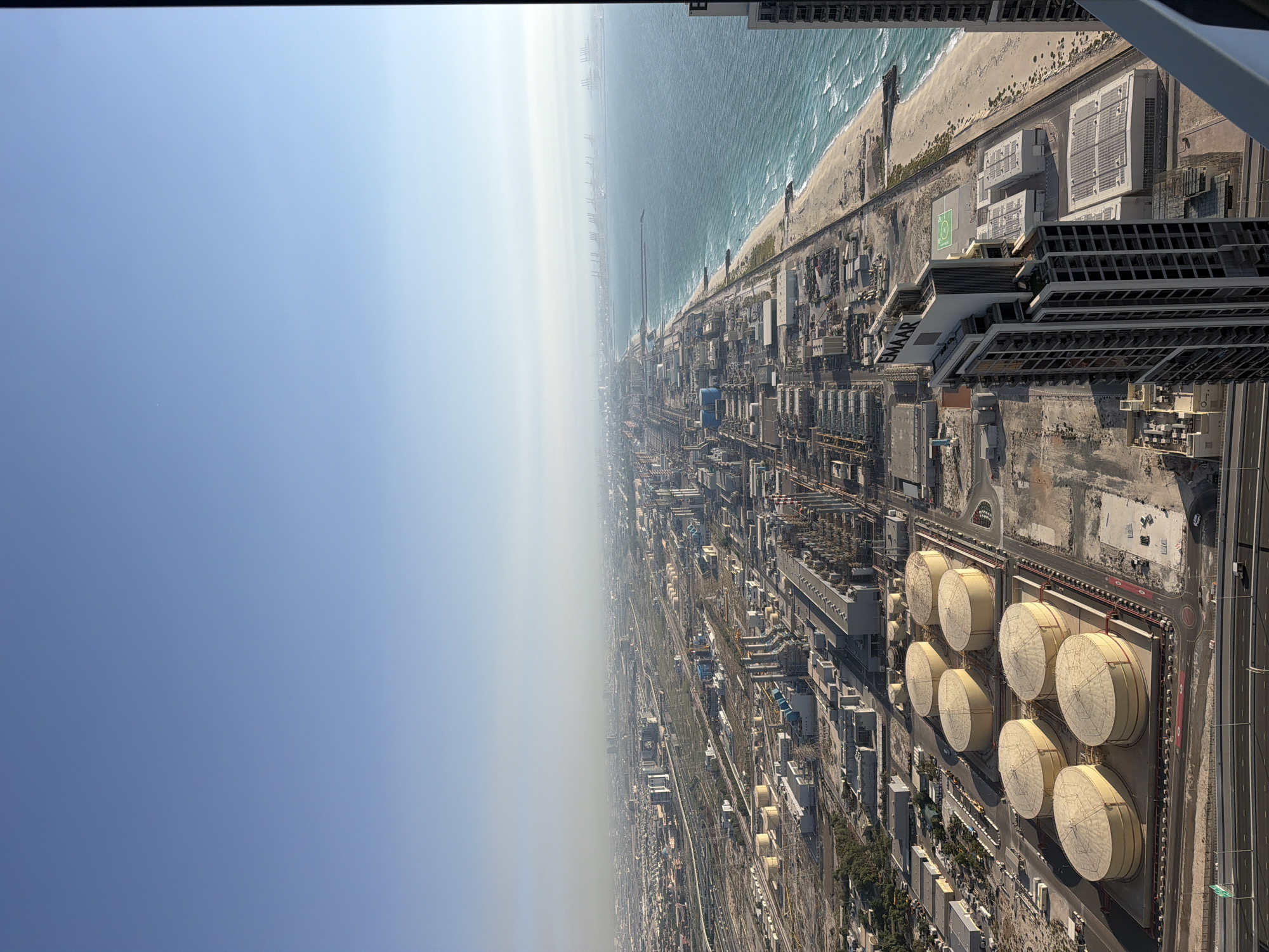 An aerial, high-angle photograph captures a sprawling coastal cityscape under a bright, clear blue sky, which transitions to a lighter, hazy tone towards the distant horizon. The scene is divided by a sandy beach where turquoise ocean waves break.
Beyond the beach, an extensive urban and industrial landscape unfolds. A prominent cluster of large, beige cylindrical storage tanks with conical roofs is visible, neatly arranged in rows within a structured compound. The land is densely packed with numerous industrial buildings, an intricate network of pipes, roads, and other infrastructure, indicative of a significant industrial and commercial hub.
Further along the distant coastline, faint structures are visible through the atmospheric haze, suggesting a large port or shipping area. In the mid-ground, a multi-story building features a green helipad marked with a white 'H' and a circle. The text "EMAAR" is also visible on the side of this building. The bright sunlight casts distinct shadows, confirming it is daytime. No specific human activity is discernible. The overall vista conveys a highly developed, modern coastal area.