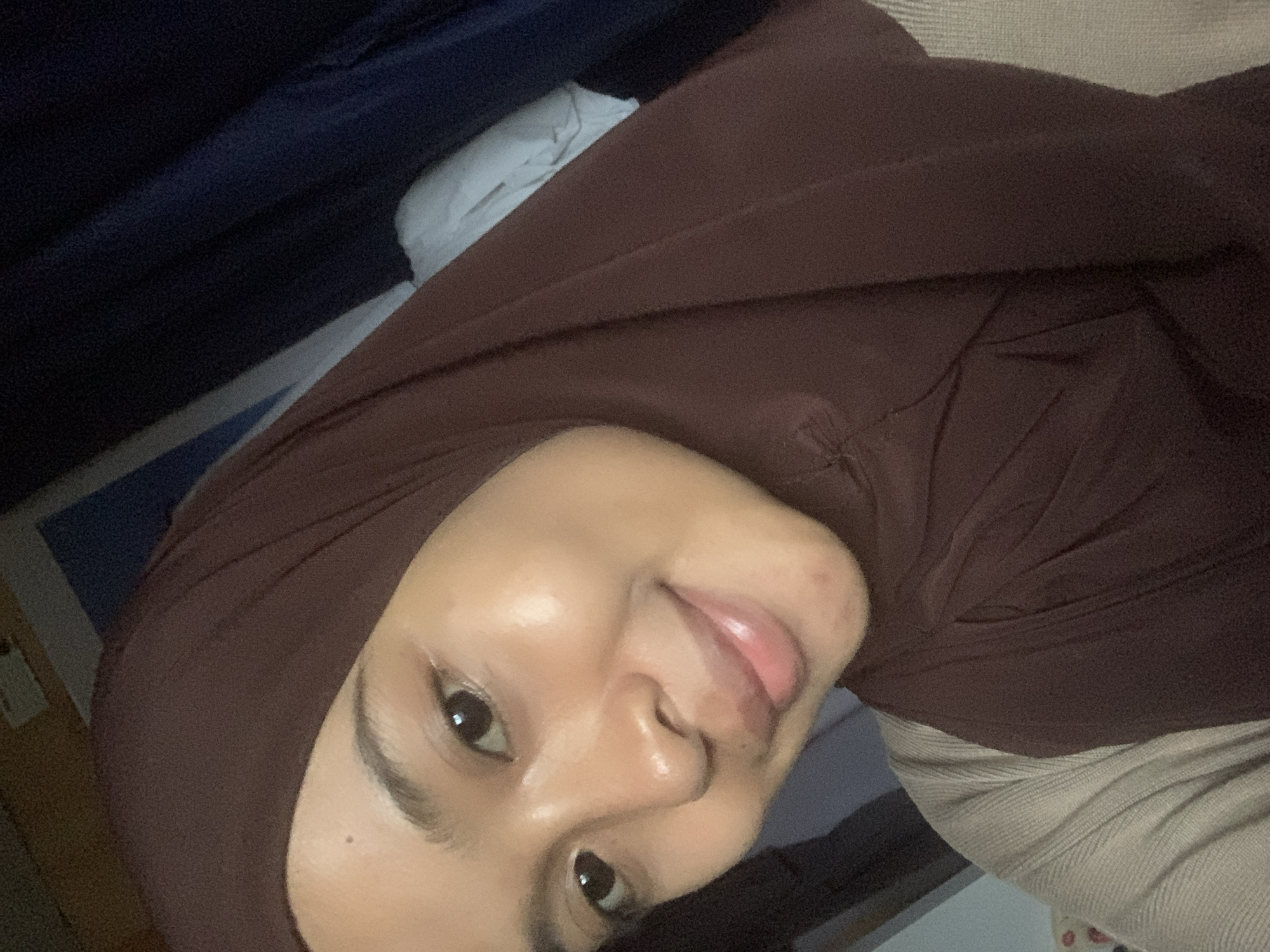 A young woman wearing a dark brown hijab and a light beige sweater is captured in a close-up, self-taken photograph. Her face is turned slightly towards the camera, and she is smiling gently. The lighting in the image highlights her dewy skin and the subtle gloss on her lips. In the background, various fabrics in shades of dark blue and a lighter blue are visible, suggesting a domestic or indoor setting. The image does not provide any specific visual clues that would allow for the determination of its location in the city of Sungai Meriam, Indonesia.