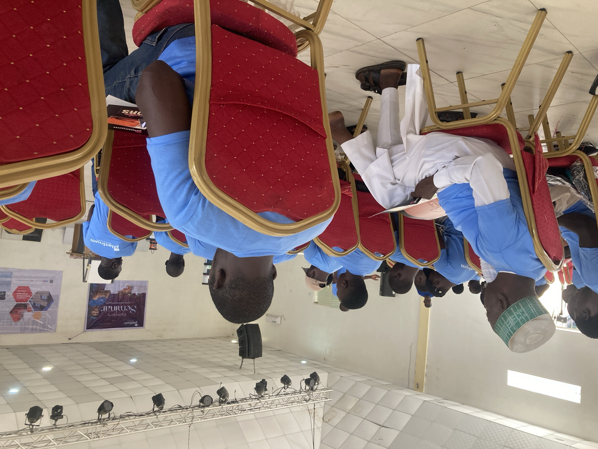 The image depicts an indoor scene, likely in a conference or seminar setting in the city of Jos, Nigeria. A row of individuals are seated on red chairs, wearing blue shirts with the word "platinum" printed on them. One individual in the row wears traditional Nigerian attire, including a white robe and a green and white hat. In the background, there's a banner advertising a "Painter's Seminar" organized by Platinum. There are several lights on a truss above the stage. The tiled flooring gives the impression of an assembly hall or a community center. There are loudspeakers behind the row of chairs.