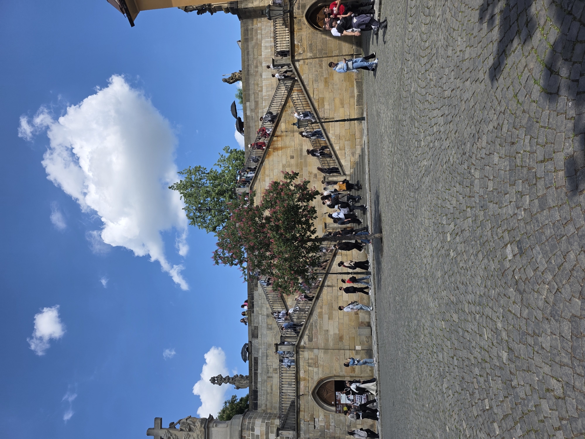 The image captures a scene on a cobblestone plaza in an unknown city. Dominating the view is a building with a staircase built into its facade, ascending in a zigzag pattern. Numerous people are populating the staircase, appearing to be tourists or locals. The sky above is blue with puffy white clouds, suggesting a sunny day. In the foreground, a cross-topped statue stands, partially obscured. A tree with pink blossoms stands near the staircase. A lamppost is visible next to the staircase.