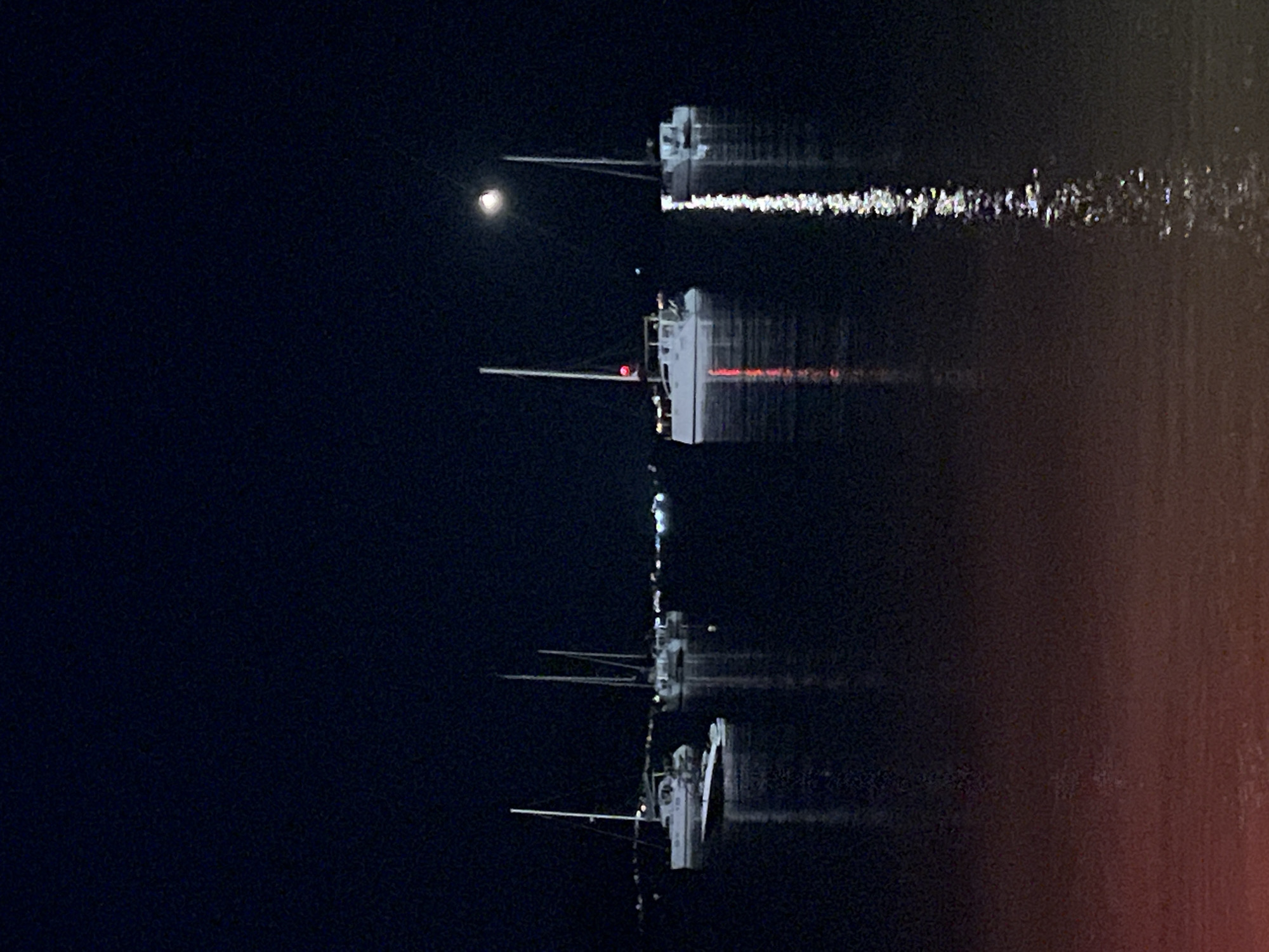 The image shows a night scene with multiple boats reflected in dark water. The sky is a deep, dark blue or black, suggesting nighttime. There is a bright, round object in the upper left quadrant, likely the moon, which is casting a soft glow. Several boats, appearing white and modern, are visible, each with what looks like a mast and some superstructure. They are positioned vertically in the frame, and their reflections are clearly seen in the water below, creating a symmetrical effect. The reflections are slightly distorted by the movement of the water, with the lights from the boats creating streaks and shimmering patterns on the surface. The lighting on the boats appears to be artificial, with some emitting red and white lights. The overall mood is serene and somewhat mysterious, highlighting the quiet beauty of boats at night. There is no visible text in the image.
