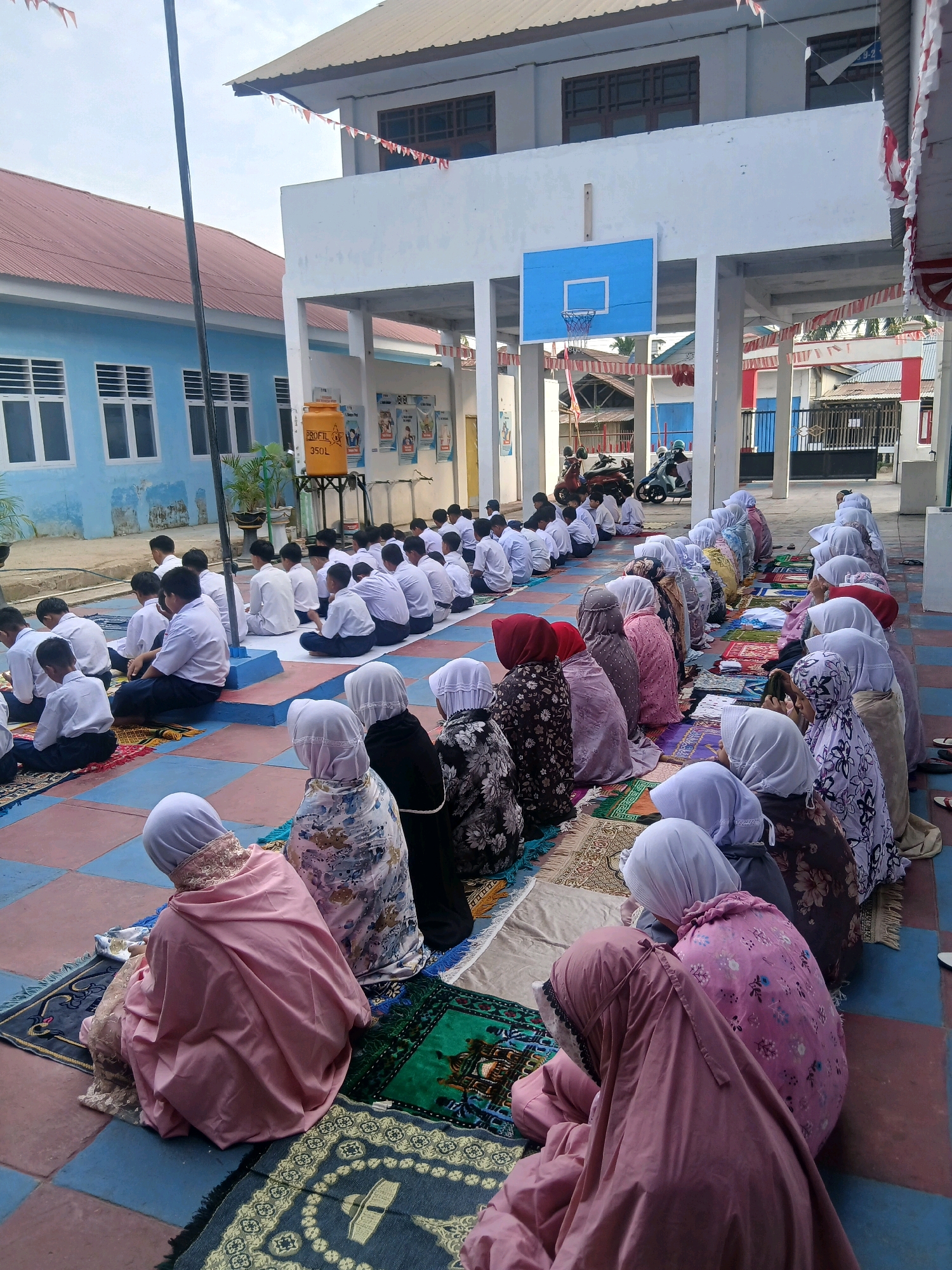 The image depicts an outdoor gathering, likely at a school in Gorontalo, Indonesia, where a group of students are engaged in prayer. The students, predominantly male, are seated on the ground in rows, facing away from the camera and towards a building. They are dressed in white shirts and dark pants, common school uniforms. A smaller group of students, appearing to be female, are also seated on prayer mats in front of the male students, facing the same direction and wearing headscarves and modest attire. The ground is paved with square tiles in alternating red and blue colors, with some areas covered by prayer mats of various designs.
In the background, a two-story building with a light blue exterior and a red-tiled roof is visible. The building has multiple windows with horizontal louvers. Another building, painted white, features a blue basketball hoop mounted on its facade. Red and white triangular flags are strung along the edge of the white building, suggesting a festive occasion or national celebration. A tall, dark flagpole stands to the left of the frame. Several motorcycles are parked in the distance, near a gate. A large yellow water tank with the inscription "PROFIL 350L" is placed on a stand near the building. The overall atmosphere appears to be one of communal worship or a school event taking place in an open-air courtyard.