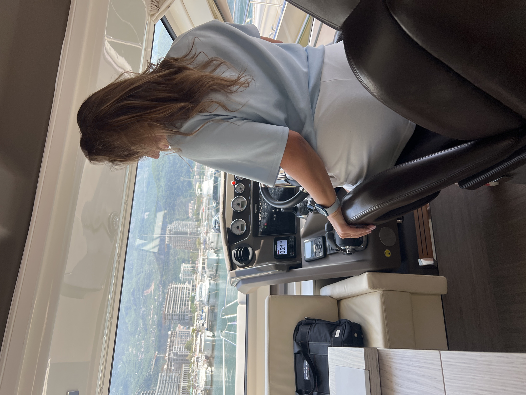 A medium shot shows a person with long brown hair sitting in the pilot's seat of a boat, facing a window overlooking the city of Budva, Montenegro. The person is wearing a light blue shirt and white pants. The pilot's seat is dark brown leather, with the person's left arm resting on the armrest. The boat's control panel is visible, featuring a steering wheel and various gauges. Through the window, the city of Budva can be seen, with a mix of residential buildings and green vegetation. In the foreground is a beige padded surface with a black bag with white text on it. The flooring appears to be dark brown wood.