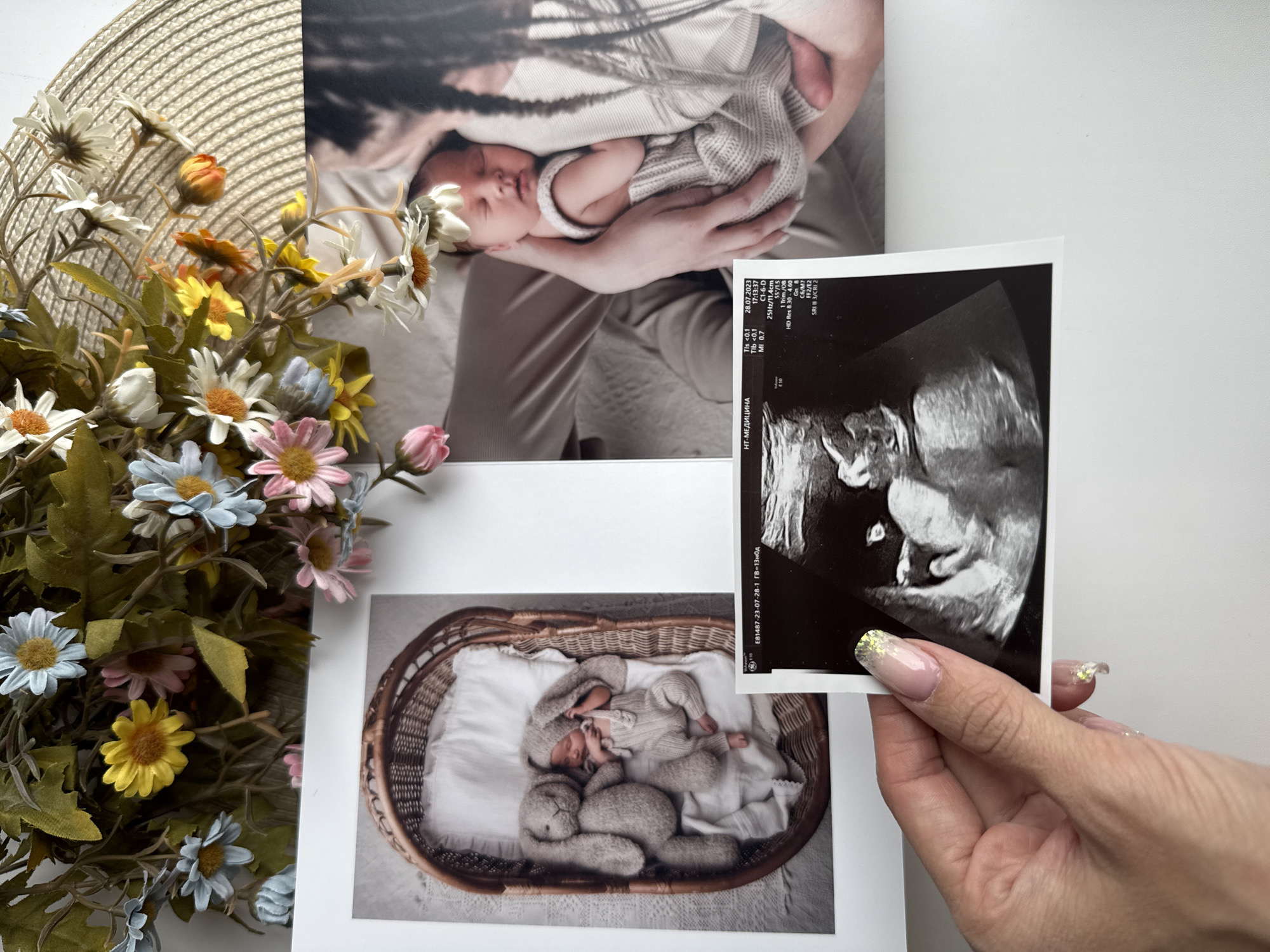 The image displays a collection of photographs arranged on a white surface, likely a table. A woman's hand, adorned with glitter-tipped nail polish, holds an ultrasound image of a fetus. Behind the ultrasound are two printed photographs of a newborn. One depicts a baby being held, and the other shows a baby in a wicker basket with a stuffed animal. A straw hat decorated with artificial flowers is positioned to the side. The ultrasound indicates it was taken at NT-Medicina on July 28, 2023. Although the OCR text indicates the Russian phrase "НТ-МЕДИЦИНА," there is no explicit indication of Rusanovo in the image.