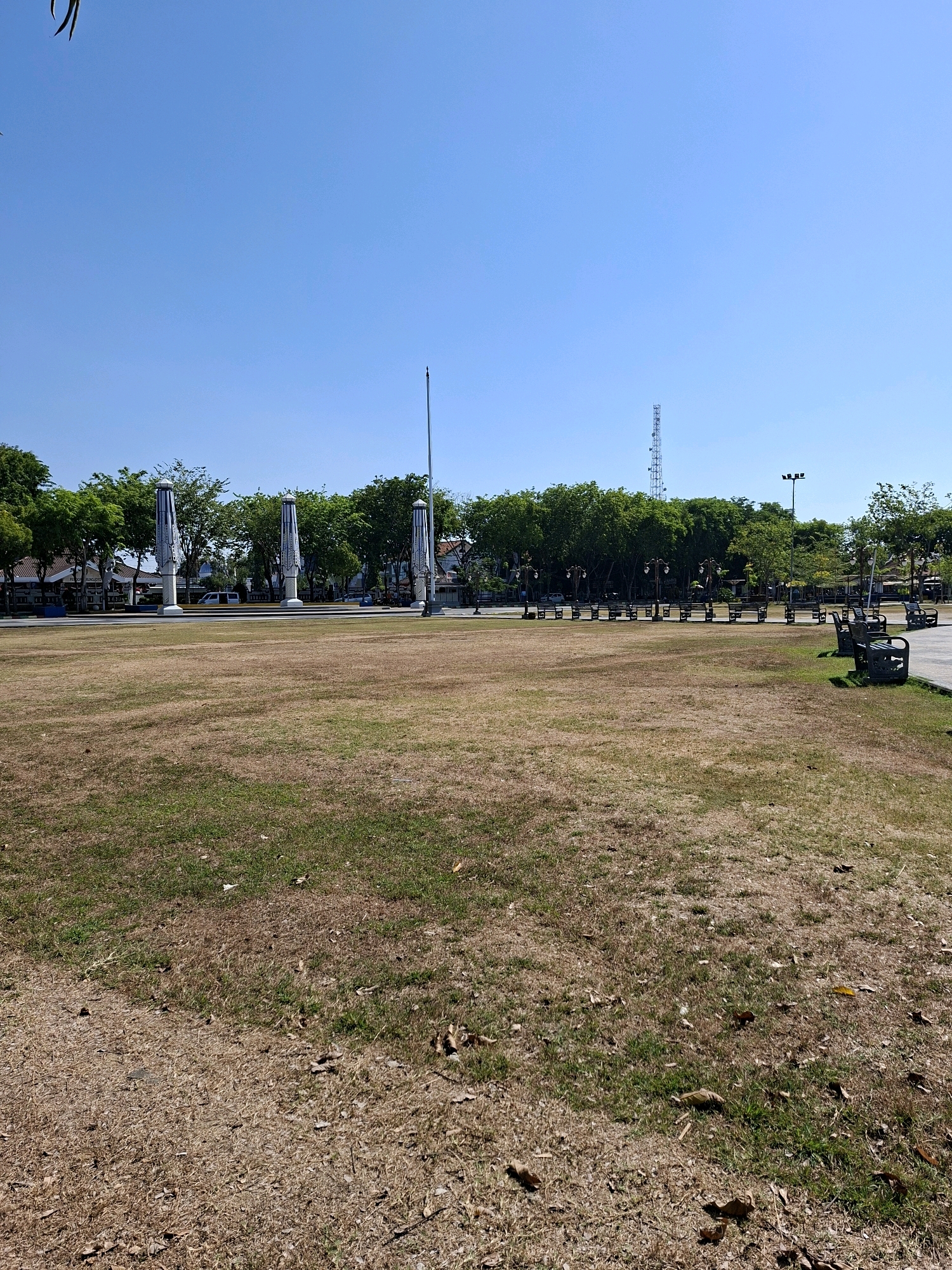 This is a photograph of a public square in Gunung Sekar, Indonesia. The square features a large, dry, grassy field with some patches of greener grass and scattered fallen leaves. In the background, there are several tall, cylindrical structures with decorative patterns and a row of trees lining the edge of the square. Beyond the trees, there are buildings and a telecommunications tower. Several park benches are visible along the perimeter of the grassy area, some of which are occupied by people. The sky is clear and blue.