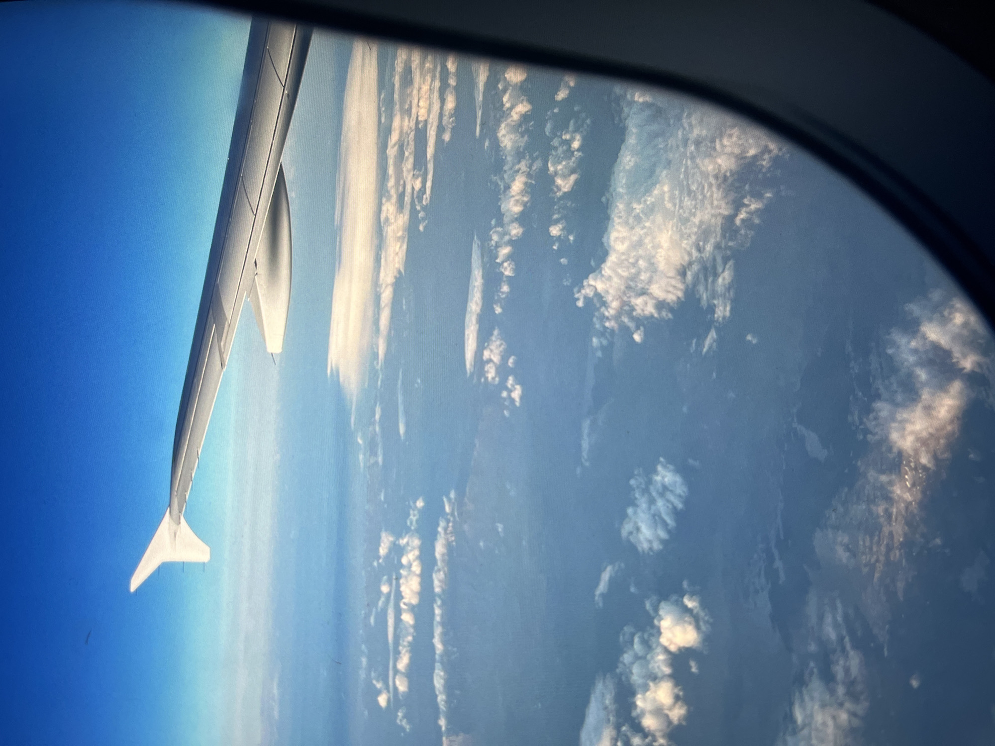 The image shows a view from an airplane window. The wing of the plane is visible on the left, extending into a clear blue sky. To the right of the wing, the view through the window reveals a sky with patchy clouds. The location is unknown.