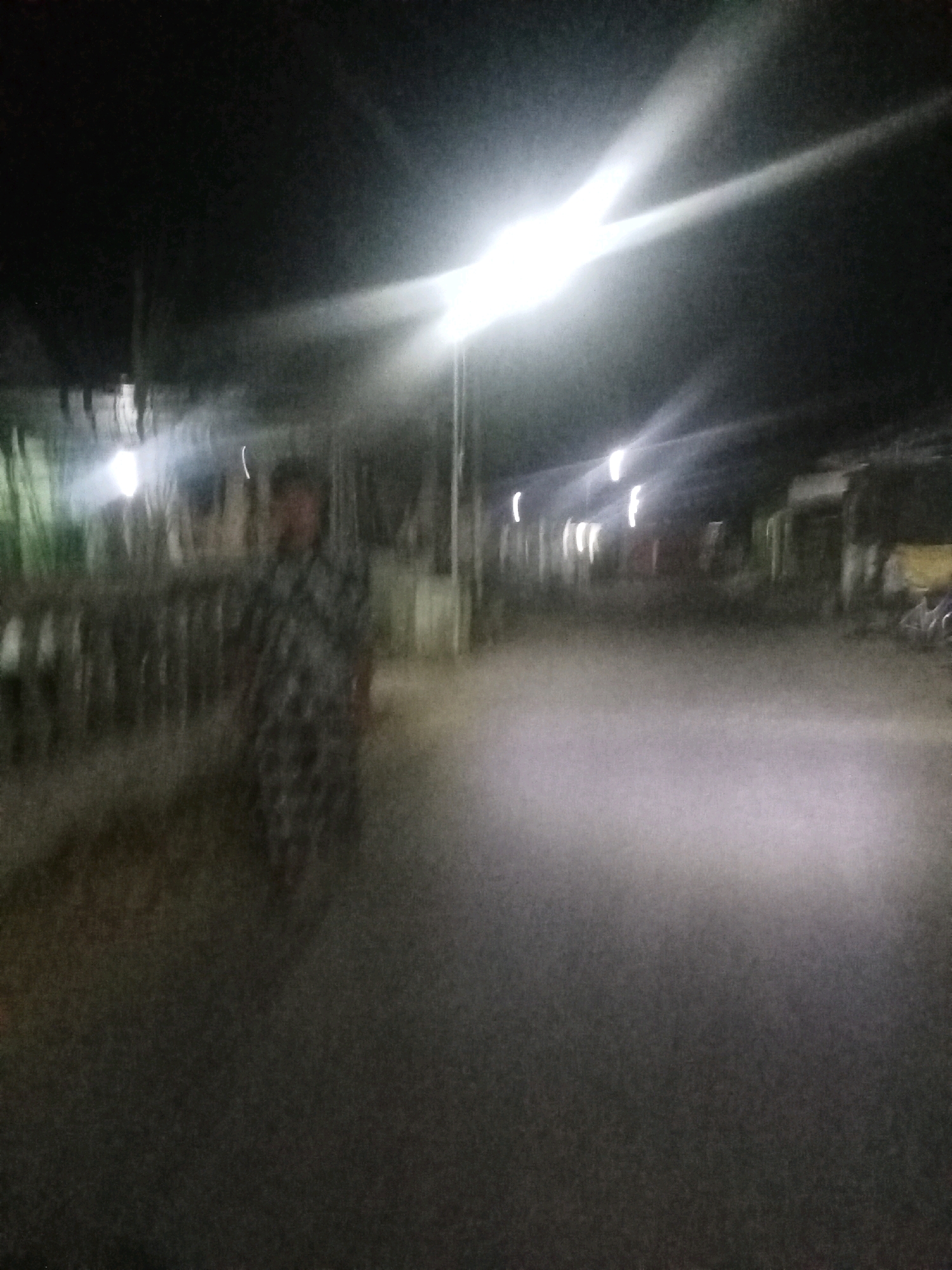 The image depicts a blurry, night-time scene of a street in Peterongan, Indonesia. A person wearing a patterned garment is visible in the mid-left of the frame, appearing to be walking or standing. Several streetlights are illuminated, casting bright streaks of light across the scene. The background shows the blurry outlines of buildings or structures lining the street, with some vegetation visible on the left. The road surface is dark and appears wet, reflecting some of the light. The overall impression is of a dimly lit urban environment at night.