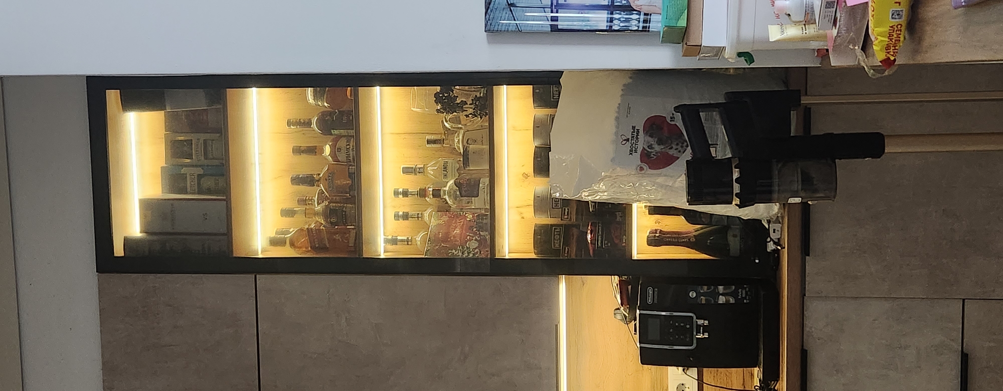 The image shows a tall, narrow display cabinet with glass shelves, illuminated by LED lights. The cabinet is filled with various bottles of alcohol and some cylindrical containers. On the left side of the image, there is a coffee machine and a portion of a grey kitchen cabinet. In the foreground, a vacuum cleaner is partially visible. The overall scene suggests a home kitchen or bar area. The city of Kazan, Russia, is mentioned as the location, but there are no specific visual cues in the image to confirm this.