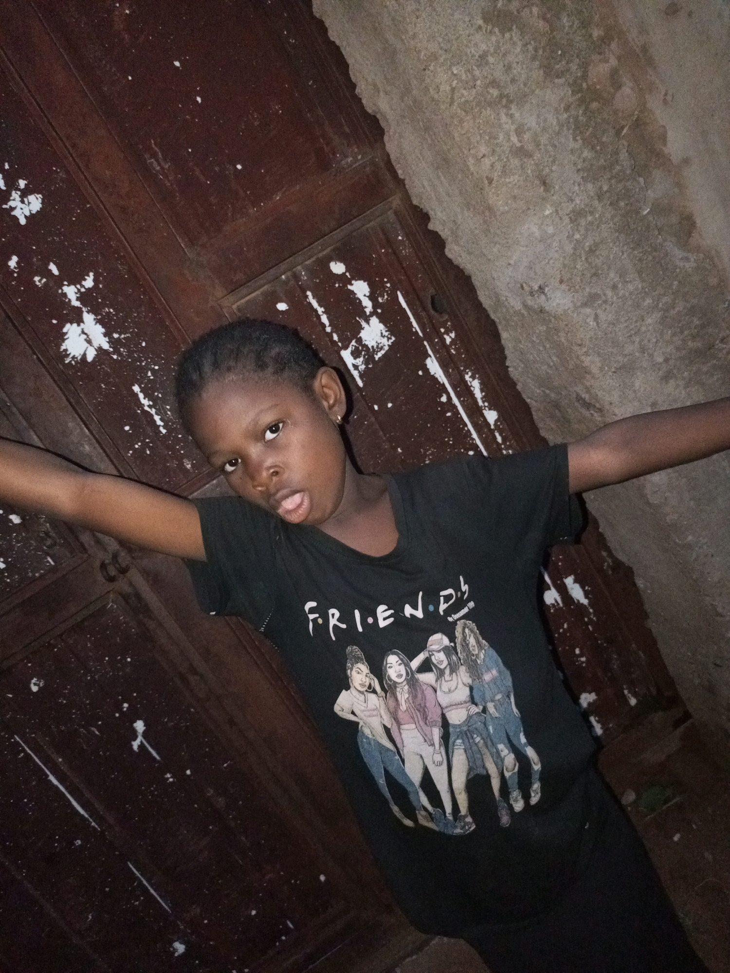 A young Black female is standing with her arms outstretched, leaning against a textured wall and a dark wooden surface. She is wearing a black t-shirt with a graphic print of five women and the word "FRIENDS" in white lettering. The background appears to be an outdoor or semi-outdoor environment, possibly an alleyway or courtyard in Tempure, Nigeria. The wall is rough and weathered, while the wooden surface looks like a door or paneling, with white splatters suggesting paint. The lighting is dim, creating shadows and highlighting the textures of the surroundings.