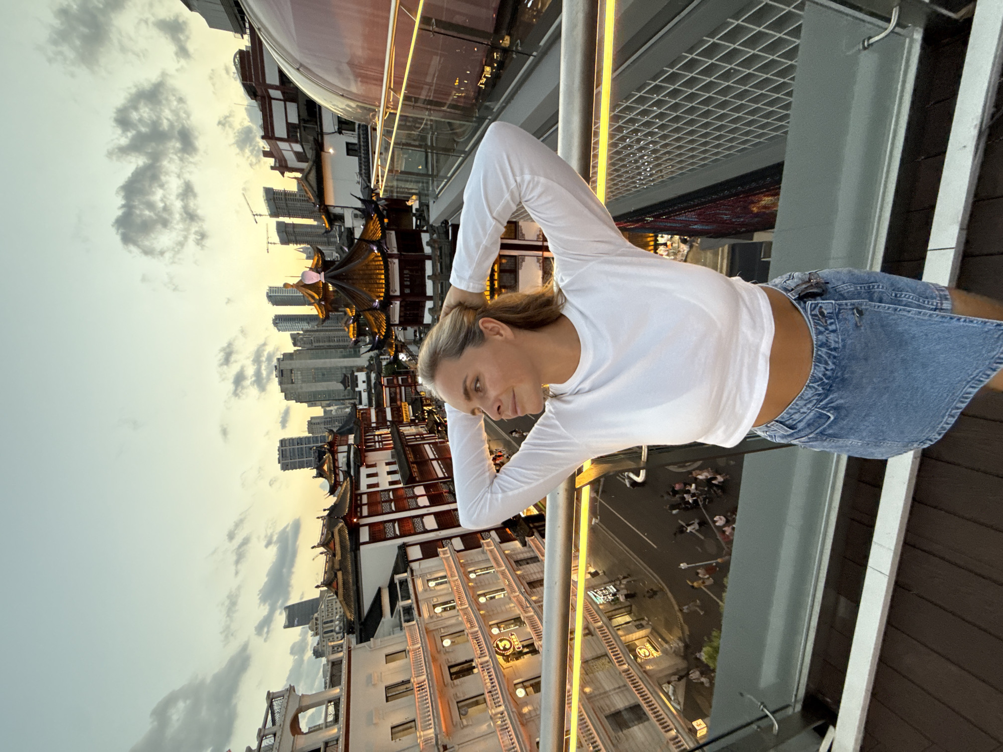 In Shanghai, China, a blonde woman with her hands behind her head is standing on a wooden deck, with a glass railing in front of her. She is wearing a white long-sleeved crop top and a denim skirt. In the background, there are several buildings with traditional Chinese architecture, featuring red and white facades with ornate roofs. Some modern skyscrapers are also visible in the distance. The sky is a pale blue with some clouds, and the setting sun casts a warm glow on the scene. Below the woman, a street is visible with people walking and some vehicles.
