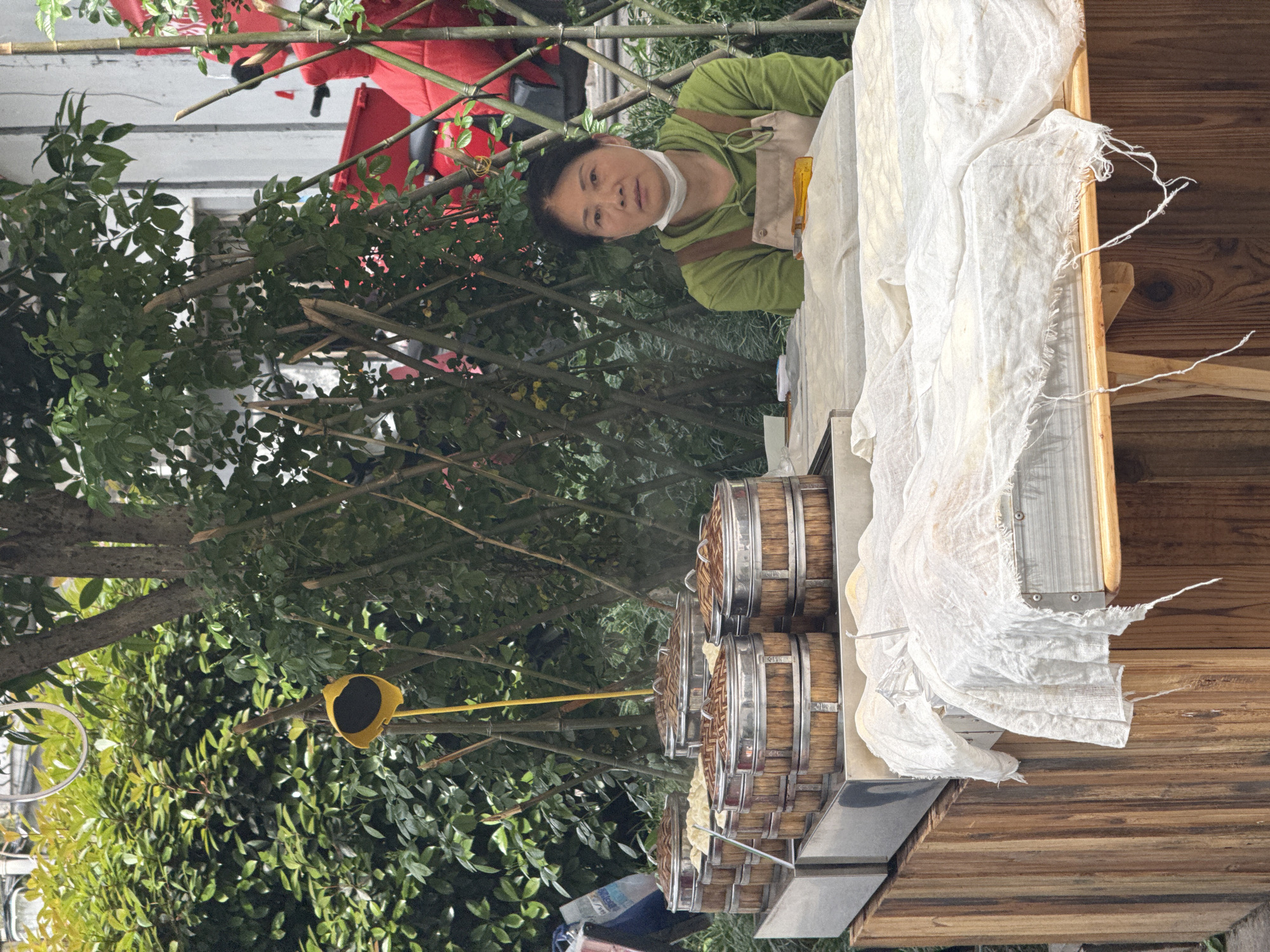 A street vendor in Yuzhong District, China, stands behind a food stall made of wood and steel. The stall has stacks of round bamboo steamer baskets. A white cloth drapes over the side of the stall. The vendor wears a green hooded sweatshirt and a light-colored apron and face covering. A yellow object hangs amongst foliage. The background includes foliage and a red object.
