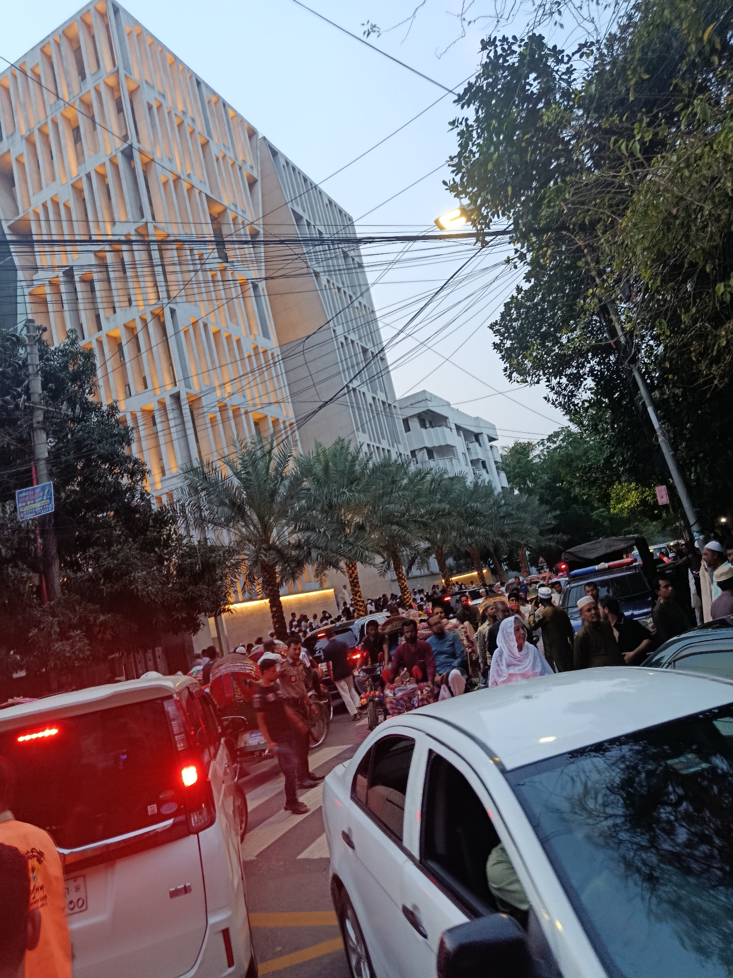 The image captures a bustling street scene in Dhaka, Bangladesh, likely during or after an event due to the presence of a large crowd of people. The crowd is composed of men, women, and children, many of whom appear to be dressed in traditional attire.
The scene is characterized by heavy traffic congestion, with several white cars and other vehicles visible in the foreground. A notable building with a modern architectural design, featuring vertical elements, stands prominently in the background. Overhead, a network of electrical wires crisscrosses the sky. Palm trees are lining the road. The image appears to have been taken at either dusk or dawn, evidenced by the lighting and sky color.