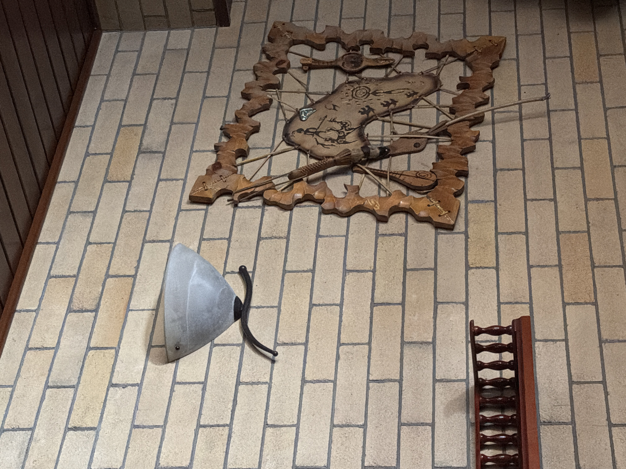 This image shows a wall with light-colored brickwork. A light fixture with a frosted glass shade is mounted on the upper left side of the wall. To the right of the light fixture, there is a decorative wooden wall hanging that appears to be in the shape of a stylized horse head within a circular frame, adorned with various carved elements and symbols. Below the light fixture, there is a small, dark red wooden shelf with balusters. The ceiling above the brickwork is made of dark wood planks. The scene is indoors, and the lighting suggests it is likely nighttime or the room is dimly lit. There are no people visible in the image.
