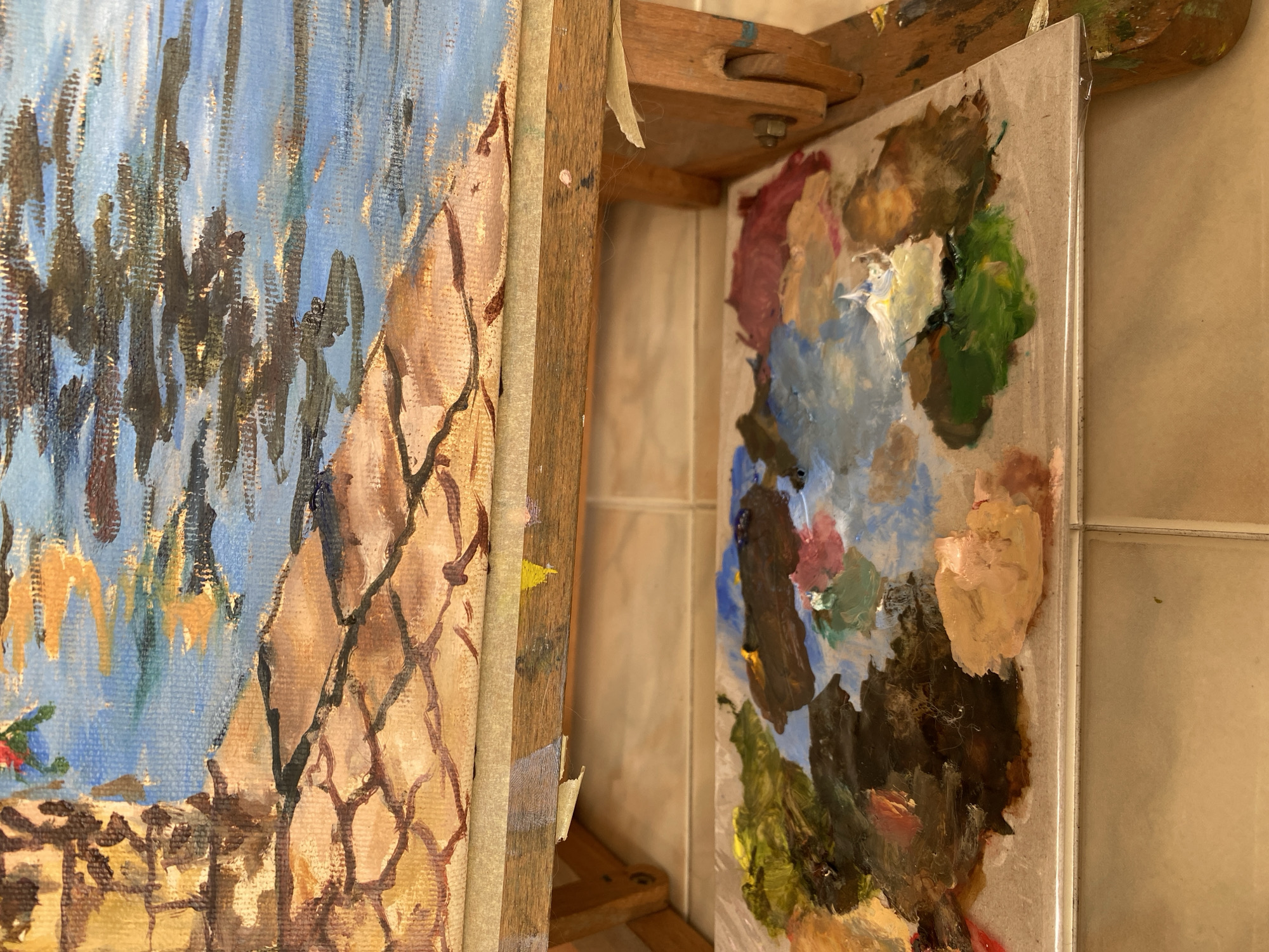 This image shows an easel holding a painting of a cobblestone path next to a body of water. Below the painting is a palette covered in various colors of oil paint. The easel is made of wood and is positioned on a tiled floor. There are no people visible in the image. The painting appears to be an outdoor scene, possibly a canal or harbor, with reflections of buildings or trees in the water. The palette is a mess of colors, indicating that the painting is in progress. The overall impression is one of an artist's workspace, with the tools of the trade laid out and a work in progress on display. The image does not contain any information or visual cues that would indicate its location in the city of Bílina, Czechia.