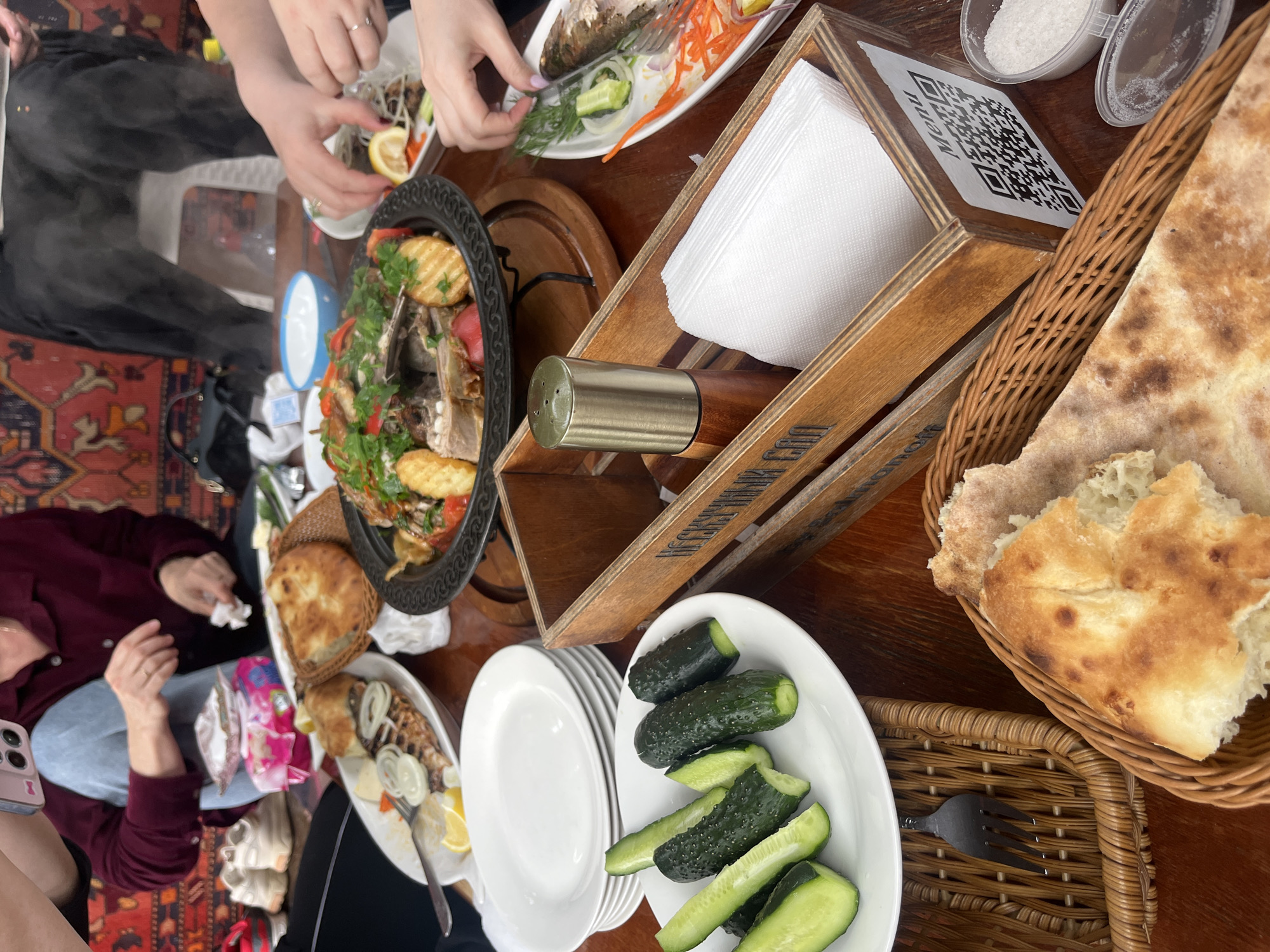 The image captures a high-angle, close-up view of a shared meal on a wooden table. Several people are seated around the table, engaged in eating and socializing. Food items include a large platter of meat and potatoes, a dish of fish, bread in a wicker basket, and a plate of sliced cucumbers. Utensils, plates, and a wooden napkin holder are visible. The QR code on the napkin holder suggests an interactive menu. The table is set on a rug. The photograph appears to be taken indoors. Based on the "Menu" visible on the napkin holder, the location may be Miatli, Russia.