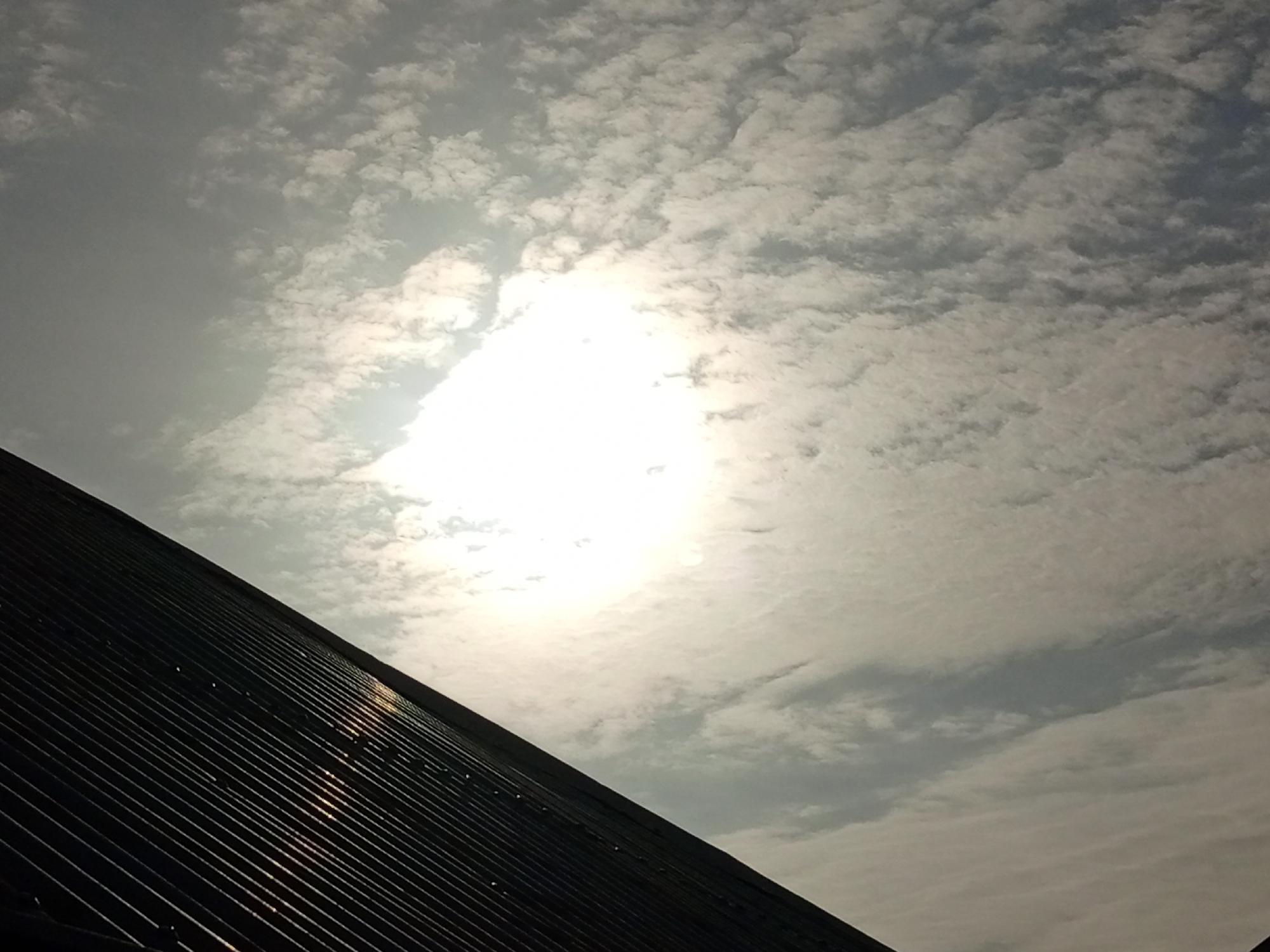 A low-angle shot captures a corrugated metal roof in the foreground, angled diagonally from the bottom left to the top right of the frame. The roof is dark, with a ribbed texture, and reflects the bright sky above. The sky is filled with a scattering of white, puffy clouds against a pale blue background. The sun is visible as a bright, diffused orb in the upper center of the image, its light washing out some of the detail in the surrounding clouds. There are no people or other objects visible in the frame. The scene appears to be an exterior shot in an urban or suburban environment, possibly Rukuba Road, Nigeria, given the context of the prompt, though no specific landmarks confirm this.