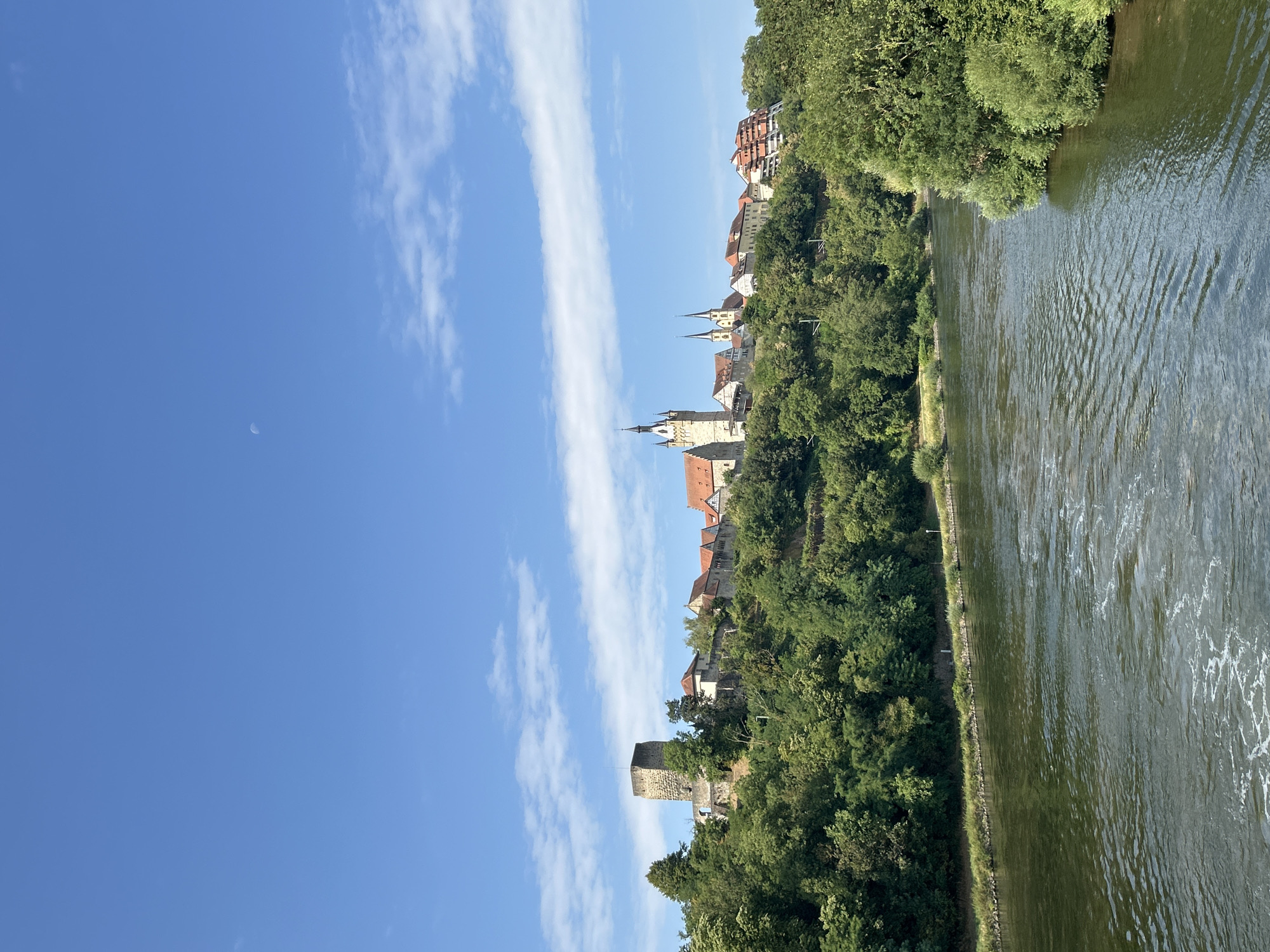 A wide, greenish-brown river flows from left to right in the foreground, with gentle ripples and some white foam visible on the surface. The riverbanks are lined with dense green trees, creating a lush, natural border. In the background, a town is situated on a hill overlooking the river. The town features several prominent buildings, including a tall, stone tower on the left, reminiscent of a castle keep. To the right of the tower, there are red-tiled roofs and other structures, including a church with a spire. Further to the right, a modern building with a distinctive tiered or stepped design is visible. The sky above is a clear, bright blue with scattered white, wispy clouds, and a faint sliver of the moon is visible in the upper portion of the frame. The overall scene depicts a picturesque view of a town nestled along a river, with a mix of historical and modern architecture set against a backdrop of vibrant green trees under a blue sky. The location is identified as the Neckar river in Offenau, Germany.