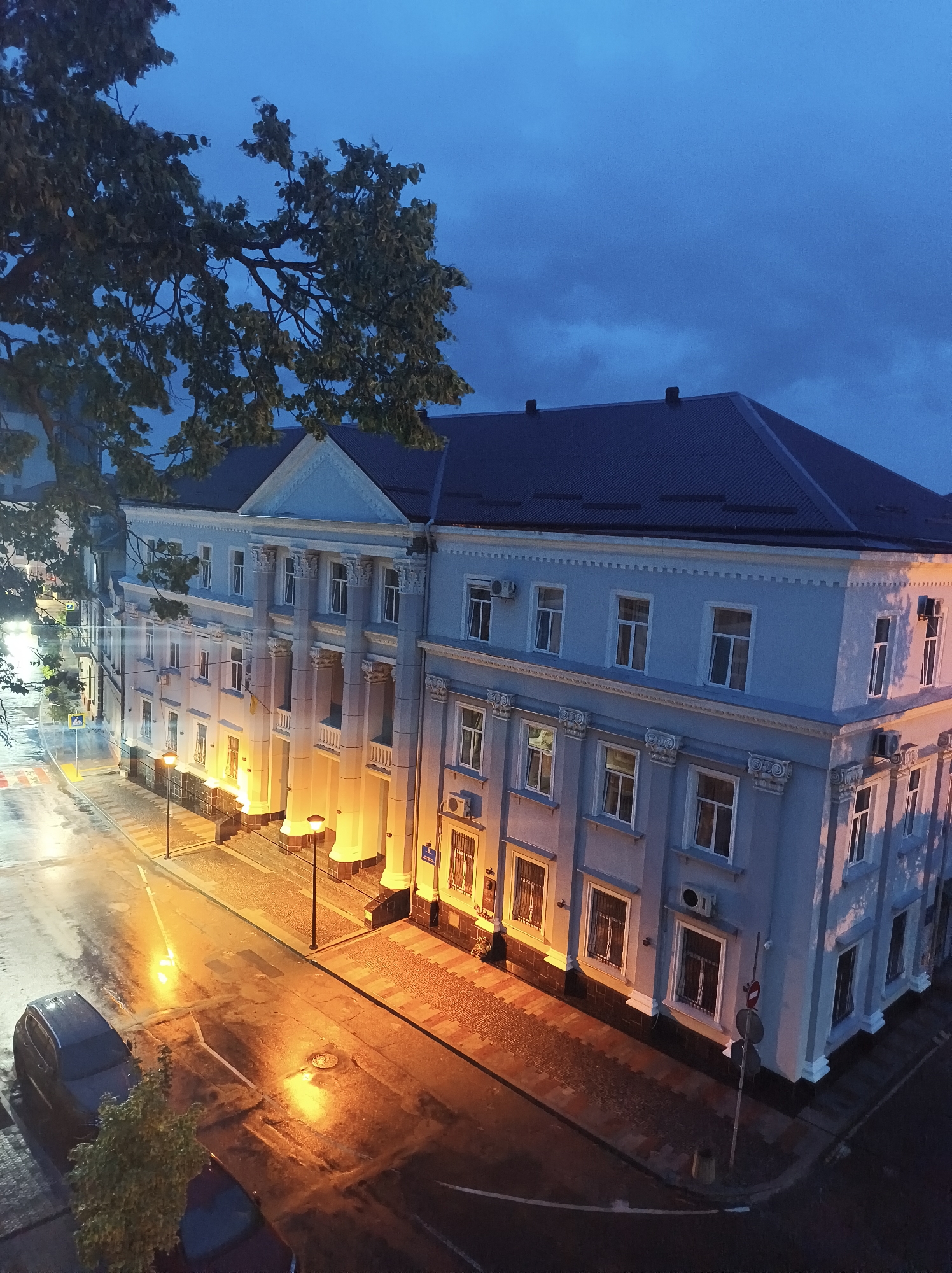 This image depicts the Ternopil Regional State Administration building at dusk. The building, a light blue, neoclassical structure with a dark roof, is illuminated by warm streetlights, casting orange glows on the wet pavement of the surrounding street. Columns line the facade, and windows are visible across its multiple stories. The sky is a deep twilight blue, with hints of clouds. A large tree with dark green leaves partially obscures the top left of the building. A dark-colored car is parked on the street to the left, and a few trees are also visible near the street. Traffic signals and streetlights dot the area. The overall impression is one of a quiet, early evening in a Ukrainian city.